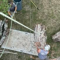 Two people working on tree branches with safety equipment. One on the ground near a board.