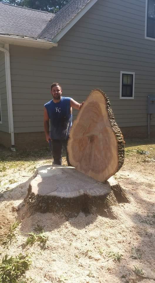 Man stands next to large tree slice, next to a stump, in front of a house.
