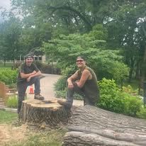 Two men in work clothes stand by cut tree, outdoors. One holds a chainsaw.
