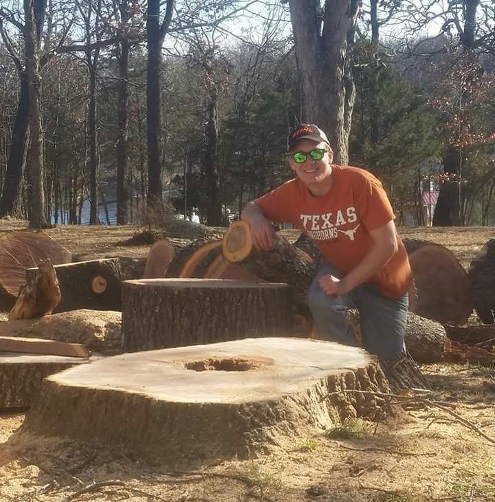 Man in orange Texas shirt kneels by a large tree stump in a sunny park.