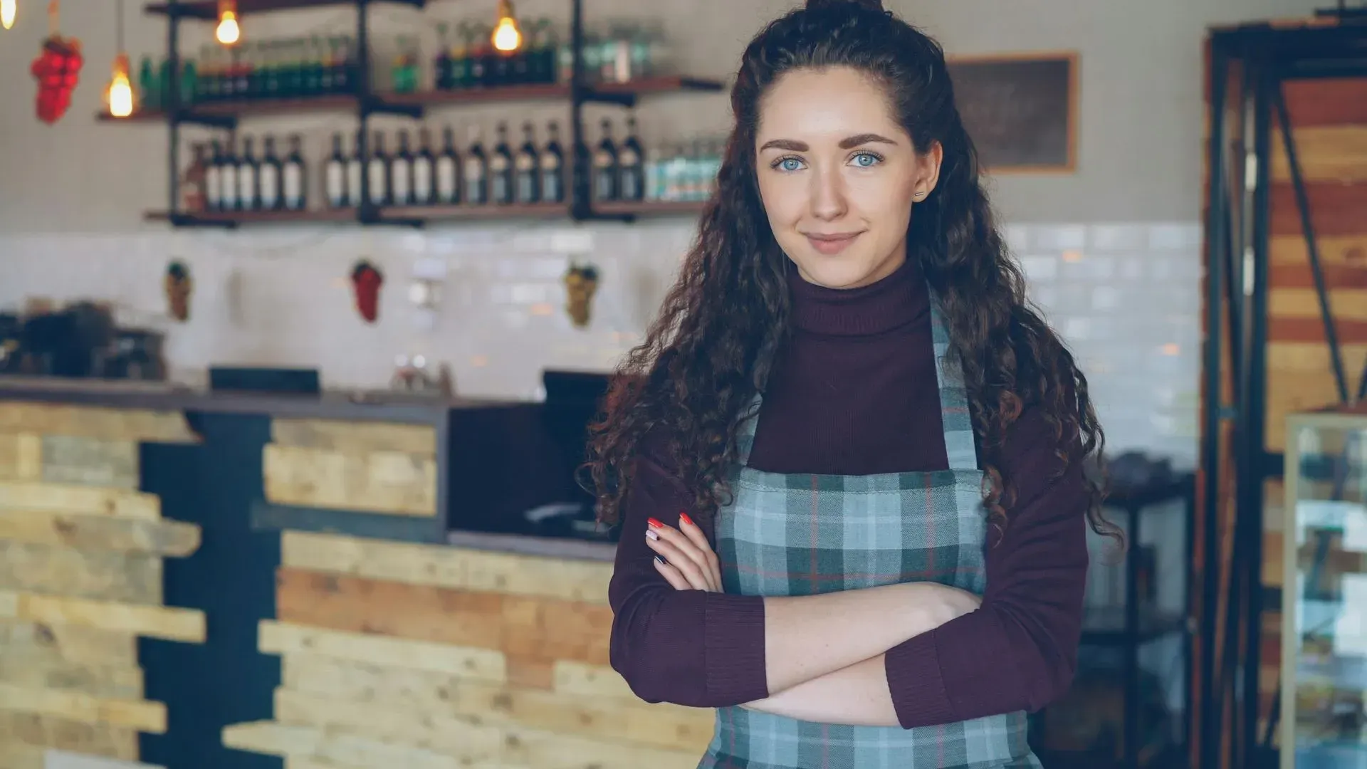 Woman standing with arms crossed in a café, wearing a plaid apron and maroon sweater.