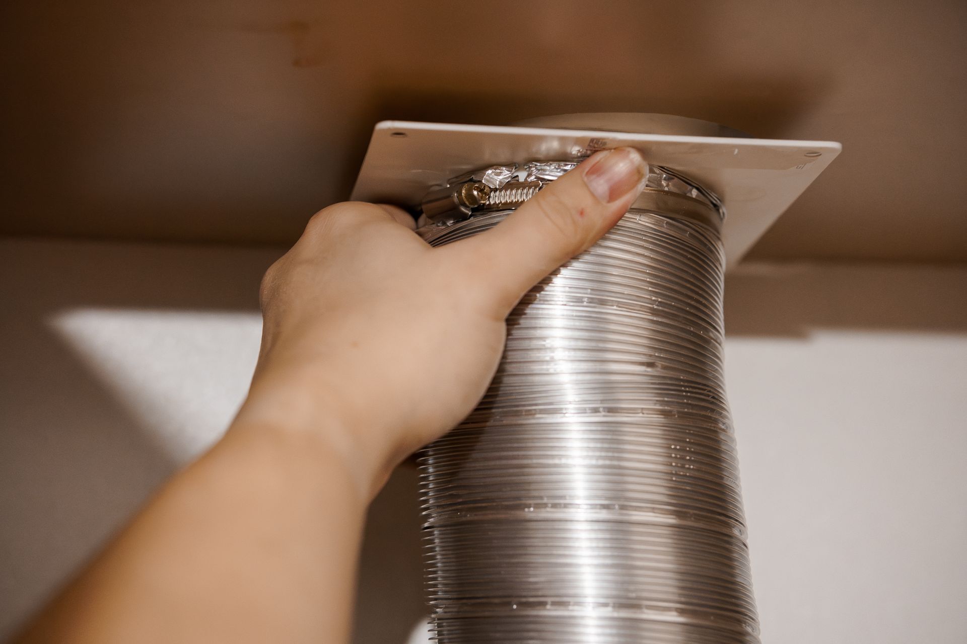 Hand holding a silver, flexible duct connected to a white square plate on a ceiling.