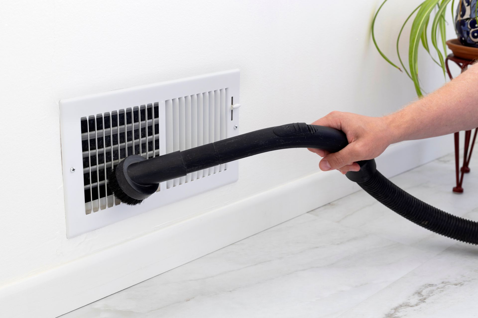 Person vacuuming a white wall vent with a black hose.