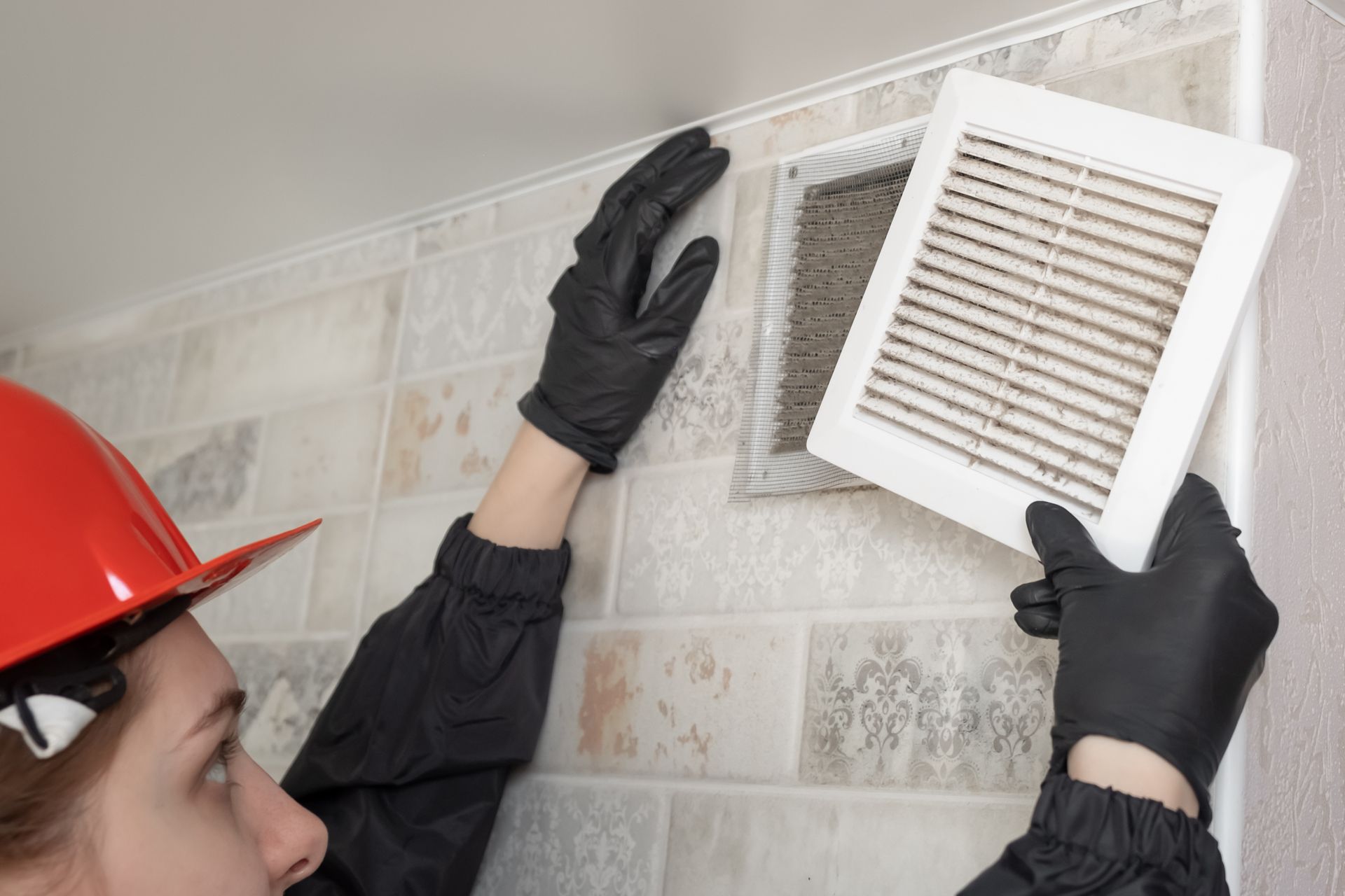 Person wearing black gloves and a red helmet removes a dirty vent cover from a wall.