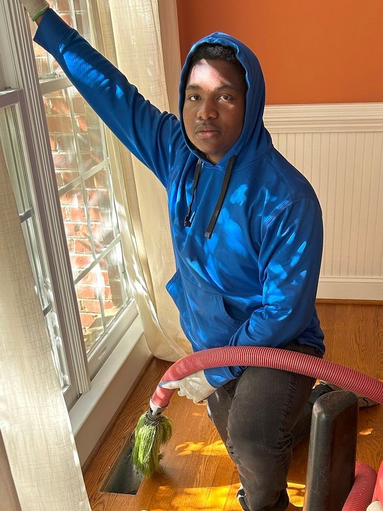 Person in blue hoodie leans near window, holding red hoop. Sunlit room, wooden trim visible. Person in blue hoodie leans near window, holding red hoop. Sunlit room, wooden trim visible.