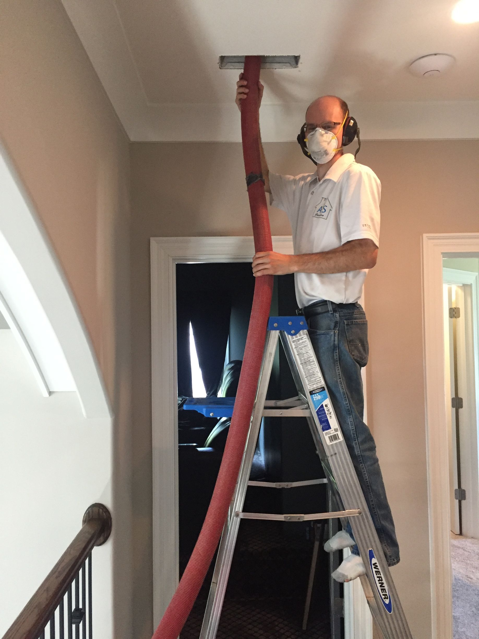 Man on ladder cleaning a vent in a hallway, using a red hose. He wears safety gear.