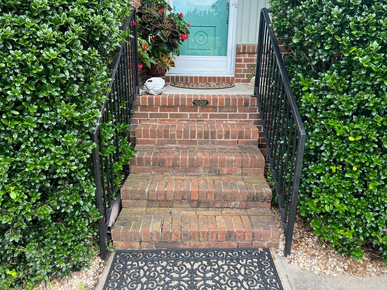 Brick steps leading to a front door with black railings, flanked by green bushes.