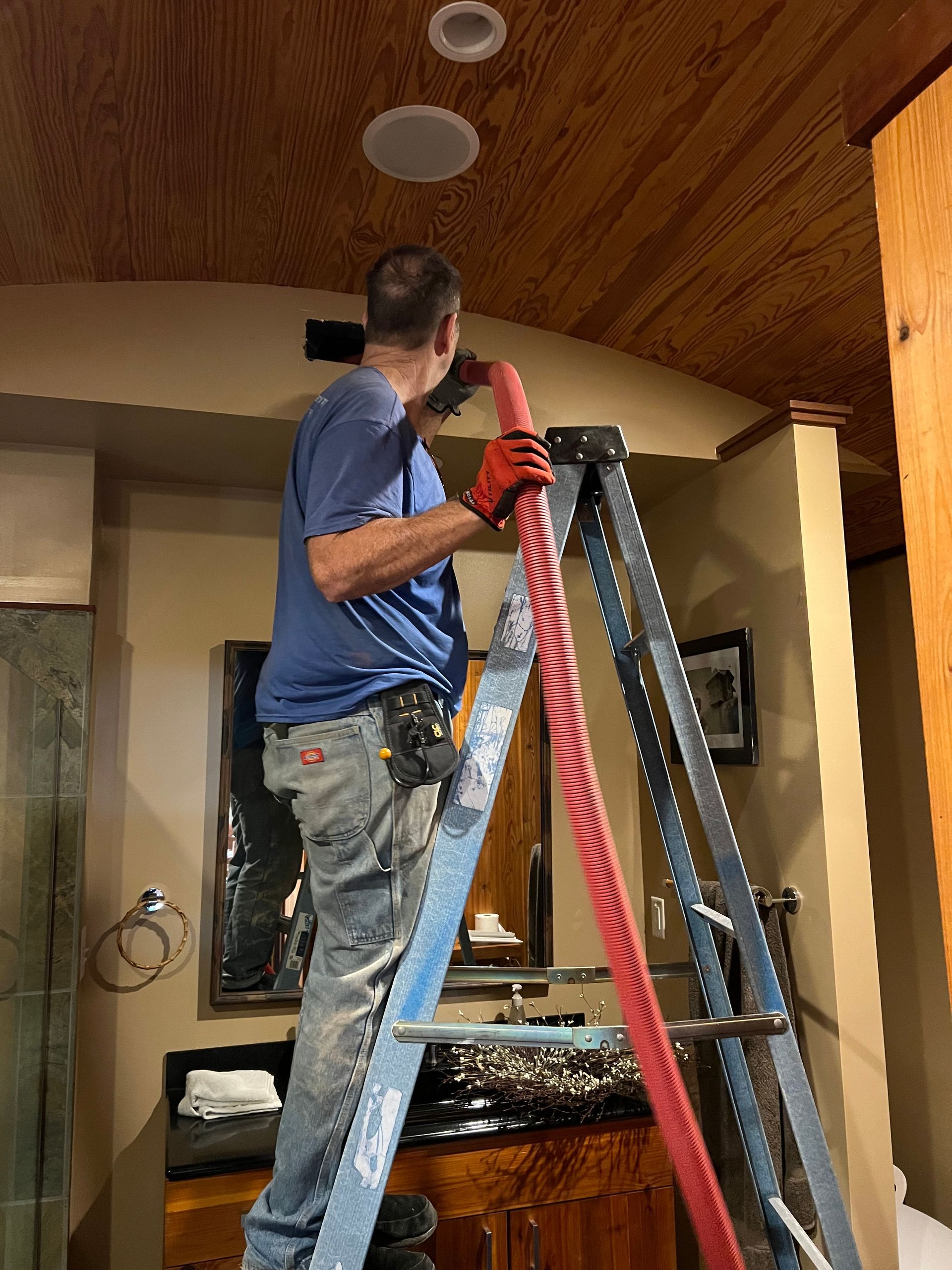 Man on a ladder working on ceiling with red hose in a bathroom.