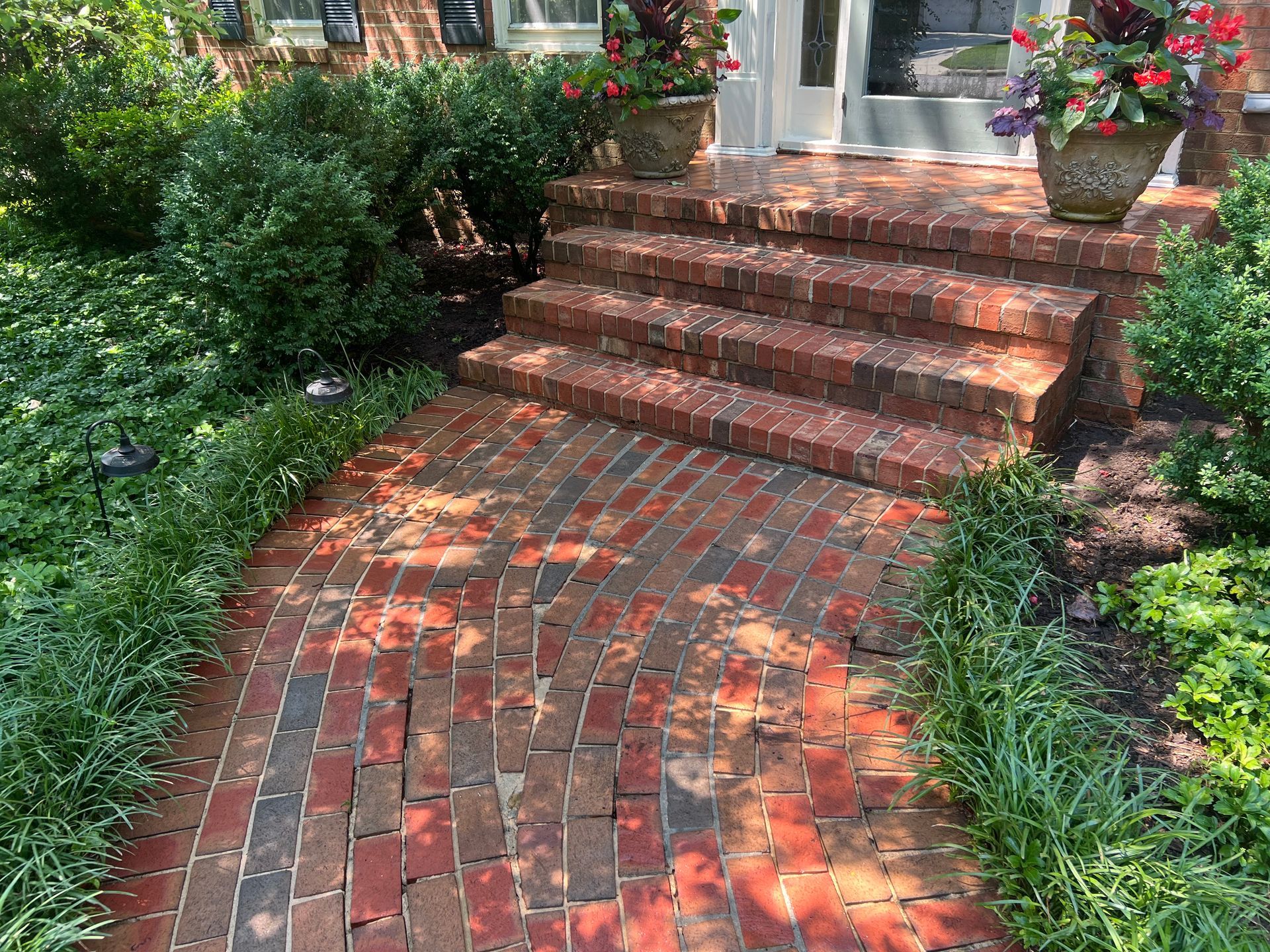 Brick walkway and steps leading to a house entrance, lined with green plants and potted flowers.
