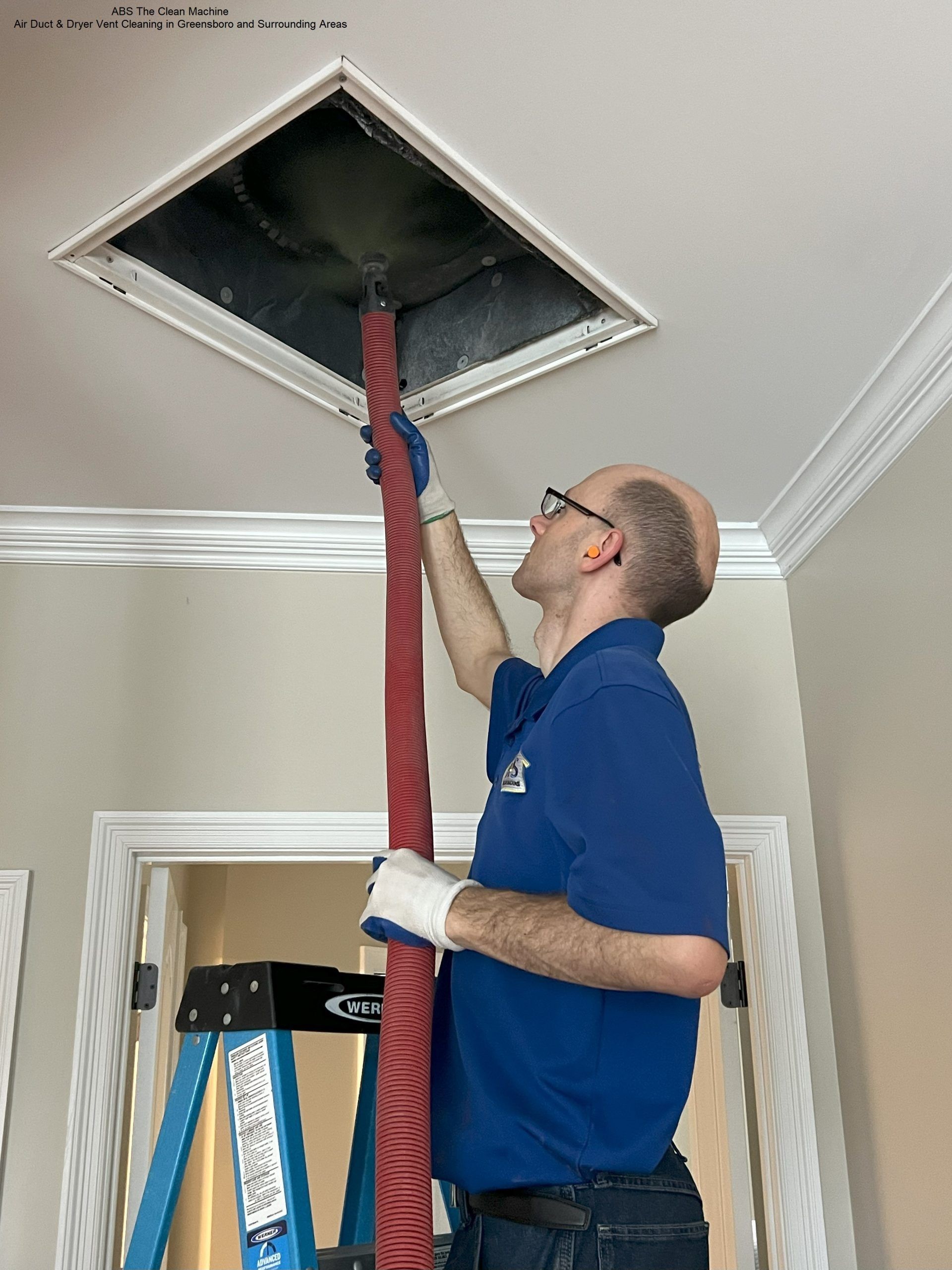 Man in blue shirt vacuums air duct from a stepladder. White gloves, red hose, white ceiling.