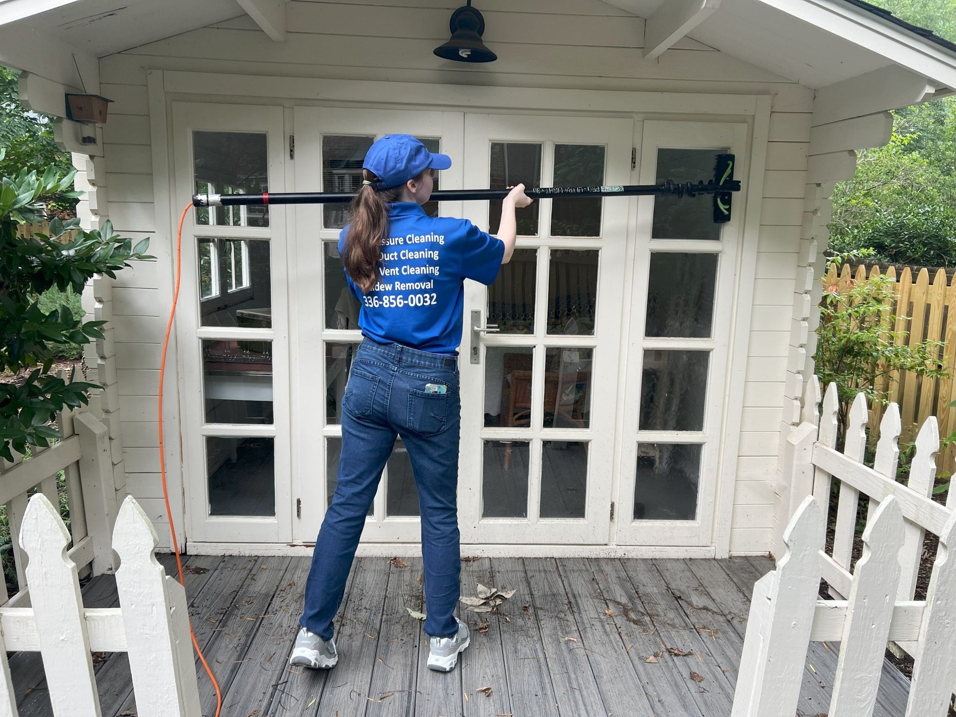 Person cleaning window, wearing a blue shirt, jeans, and a cap. Standing on a porch with white picket fence.