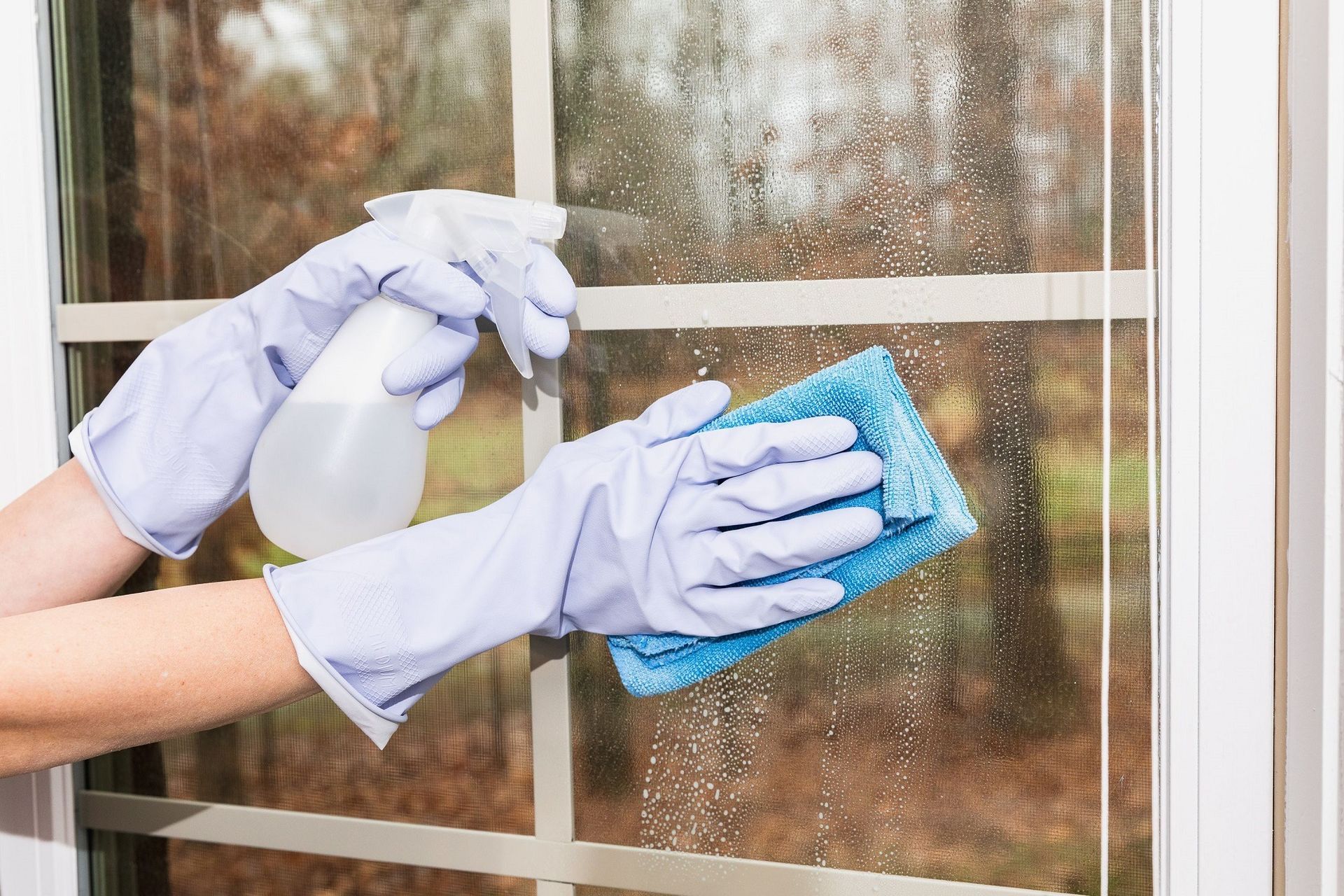 Hands in blue gloves spraying and wiping a window with a spray bottle and blue cloth.