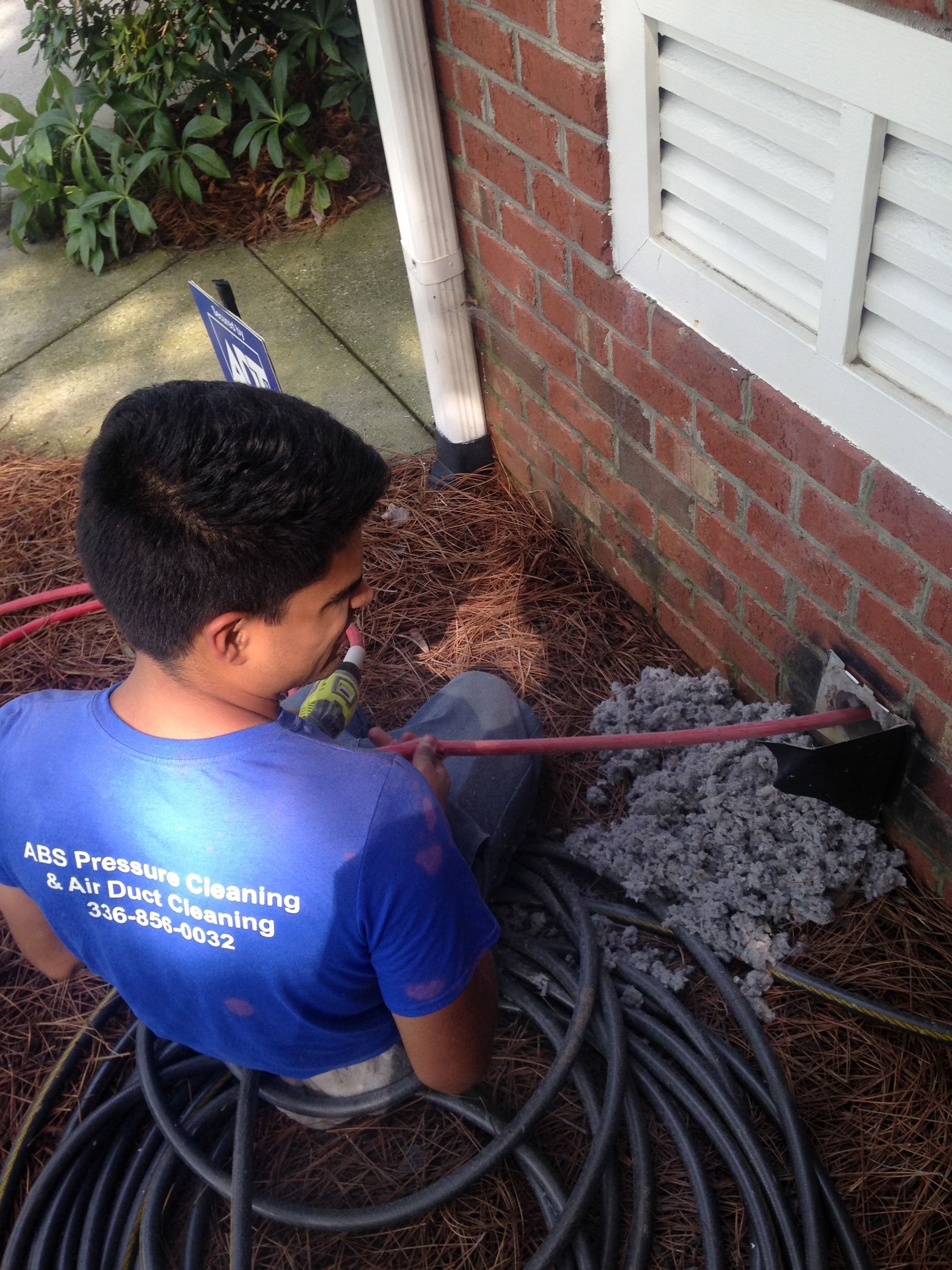 Man in blue shirt cleaning near a brick building. He is using a tool to remove debris from a vent.