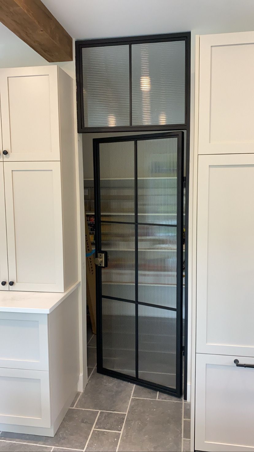 Porte vitrée à cadre noir, ouverte, donnant sur le garde-manger, placée entre des armoires blanches, sous une imposte en verre texturé.