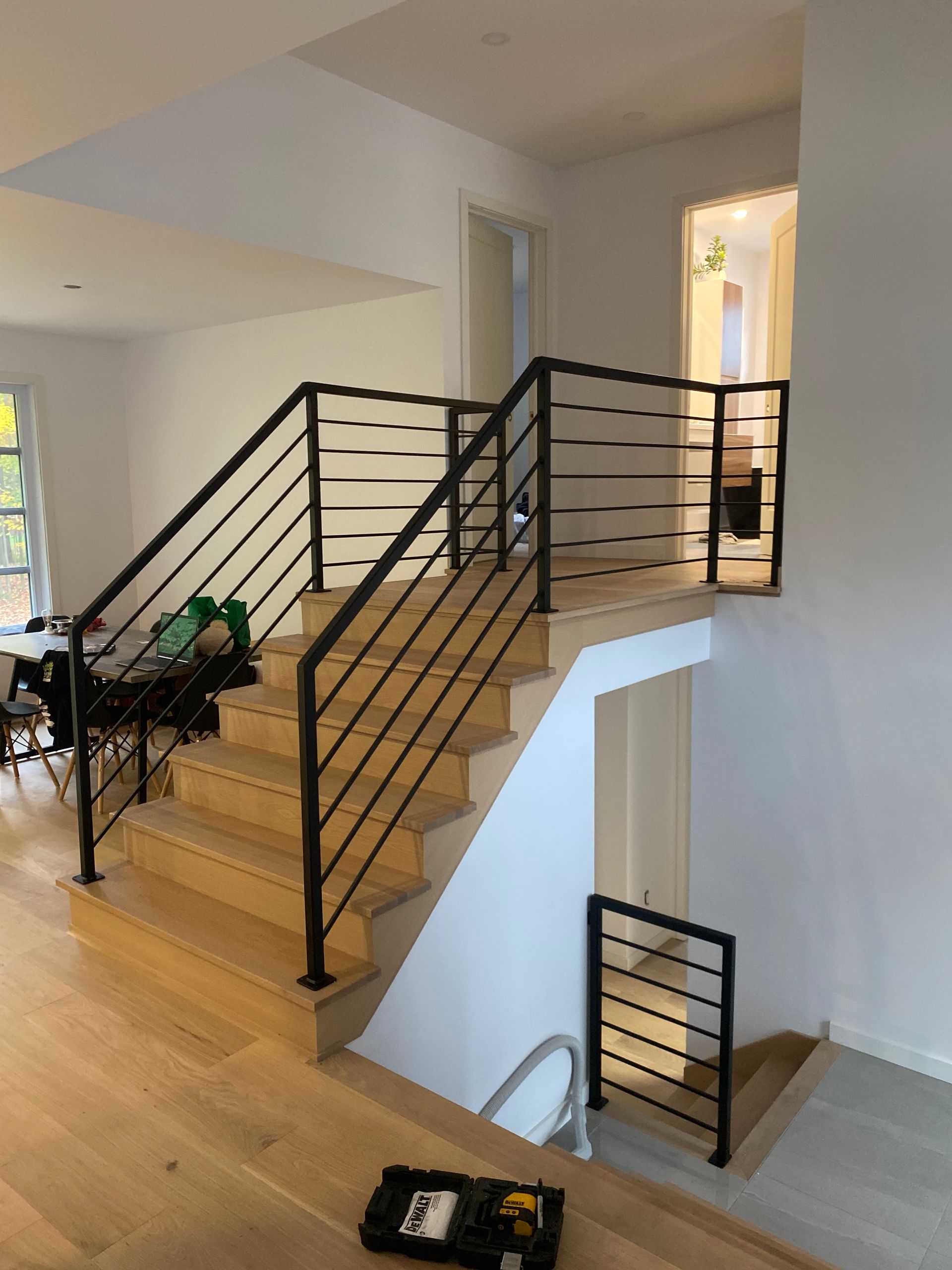 Un escalier avec des rampes en métal noir, des marches et un sol en bois clair, et des murs blancs.