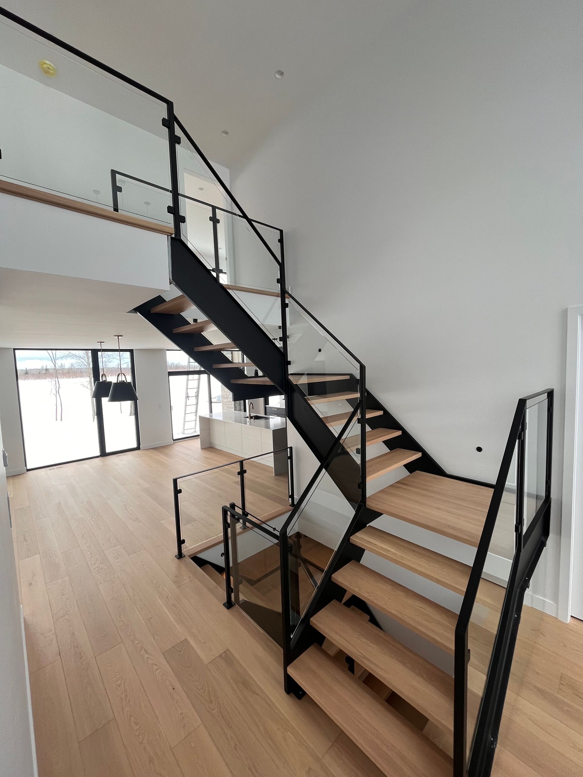 Escalier moderne avec structure en métal noir, panneaux de verre et marches en bois, dans un espace lumineux et ouvert.