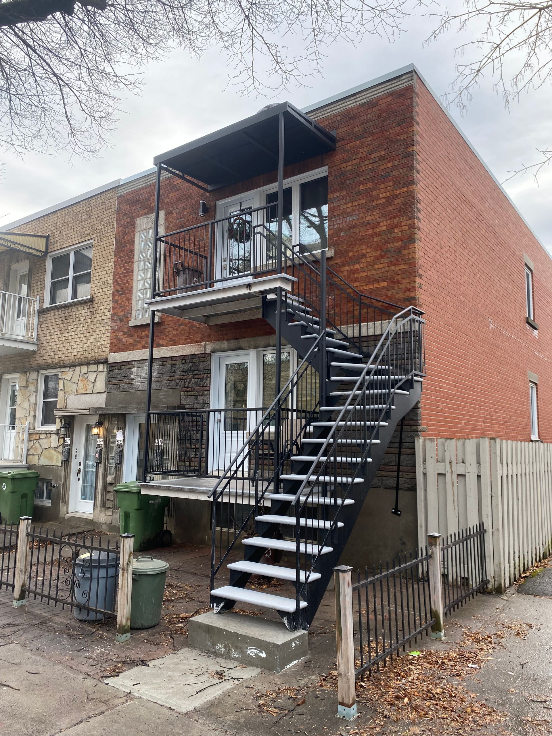 Bâtiment en briques avec un escalier extérieur en métal menant à un balcon. Des poubelles vertes sont installées près du trottoir.