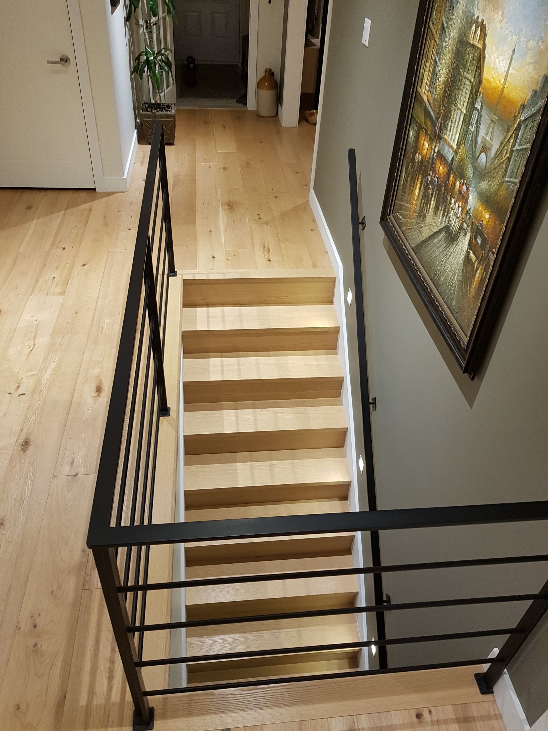 Escalier avec marches en bois et rampes en métal noir, menant à un couloir au sol clair.