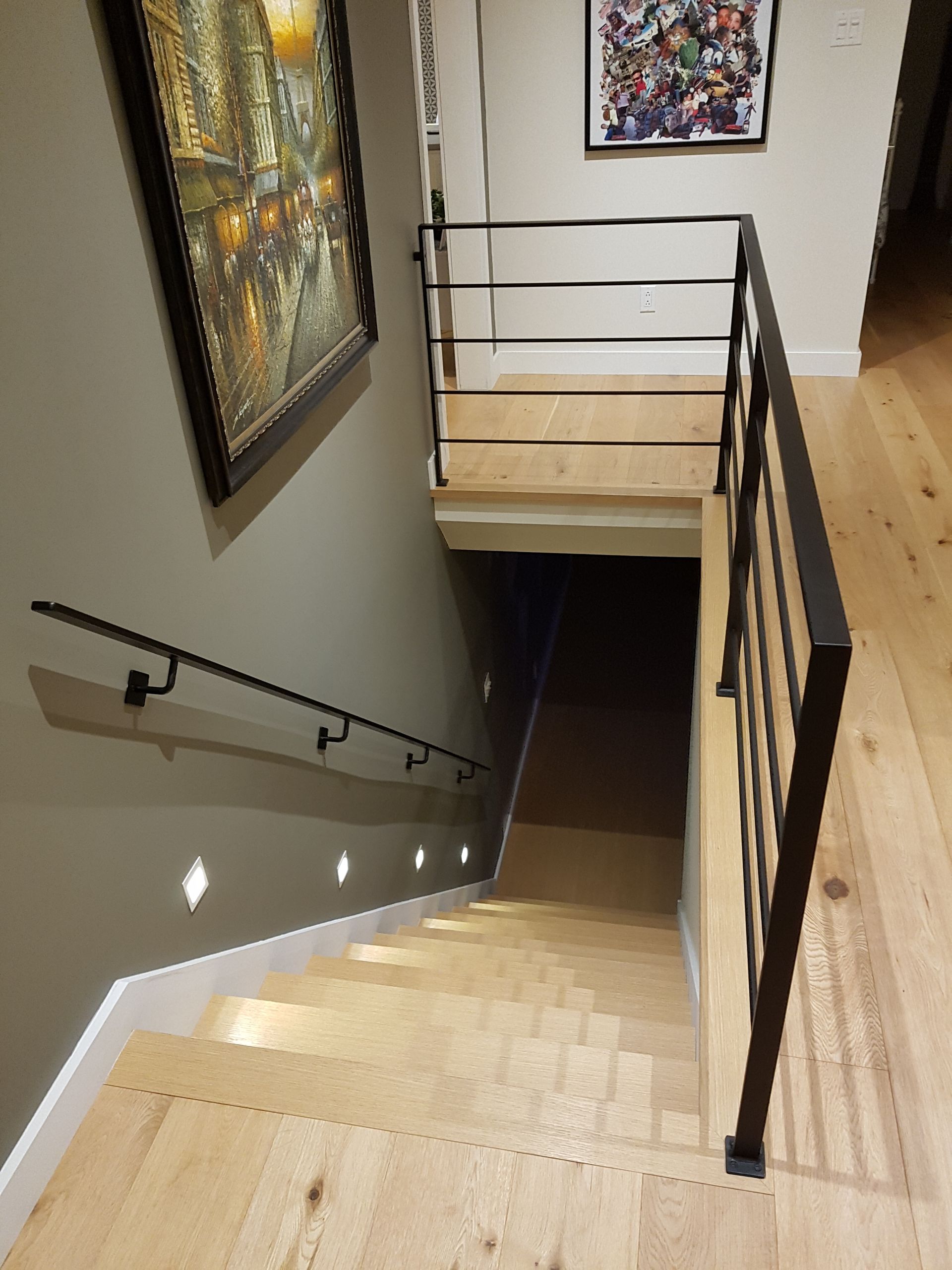 Escalier en bois menant à un sous-sol sombre, rampes en métal noir et œuvres d'art au mur.