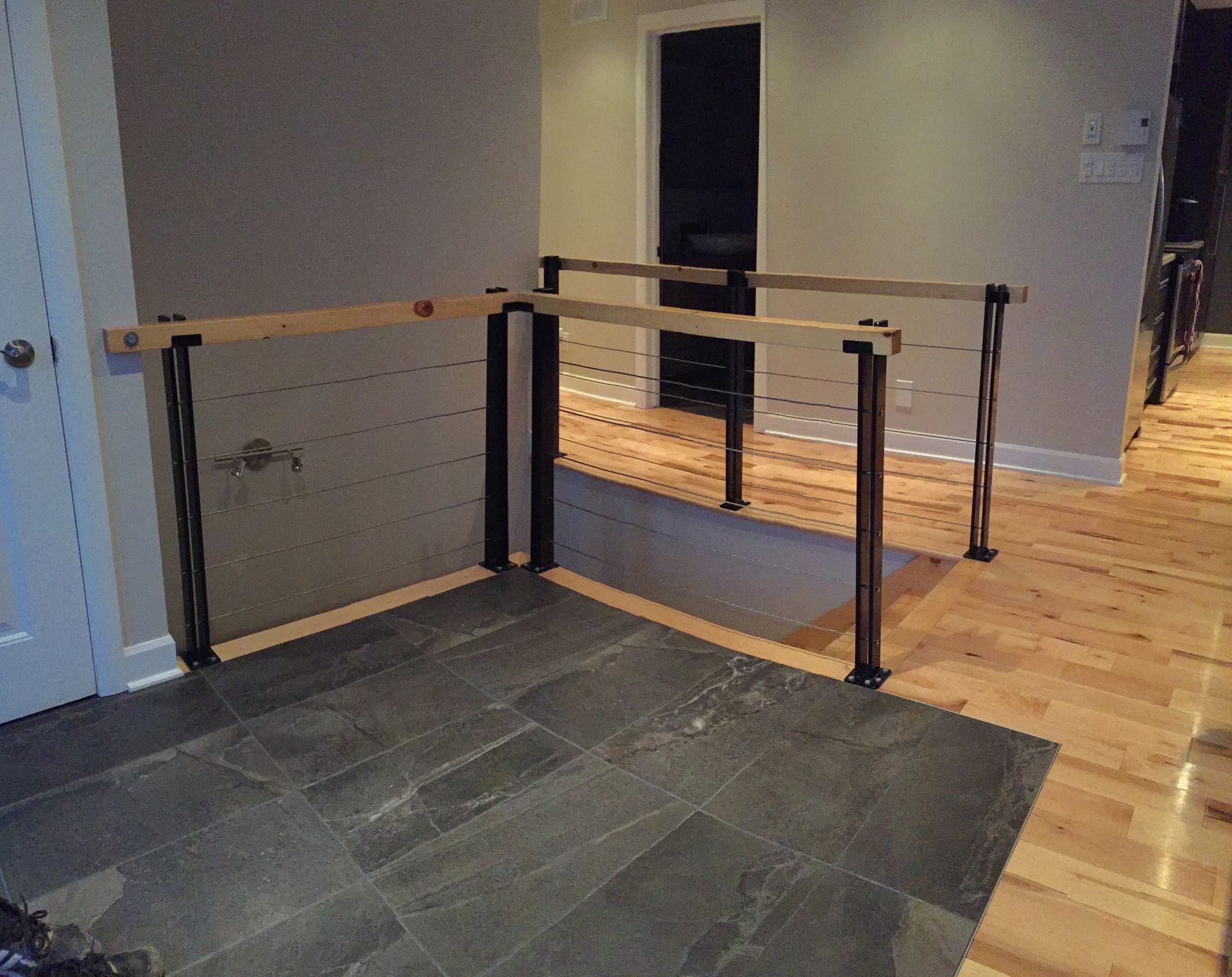 Garde-corps intérieur moderne en bois et métal noir, câbles et sol en pierre.