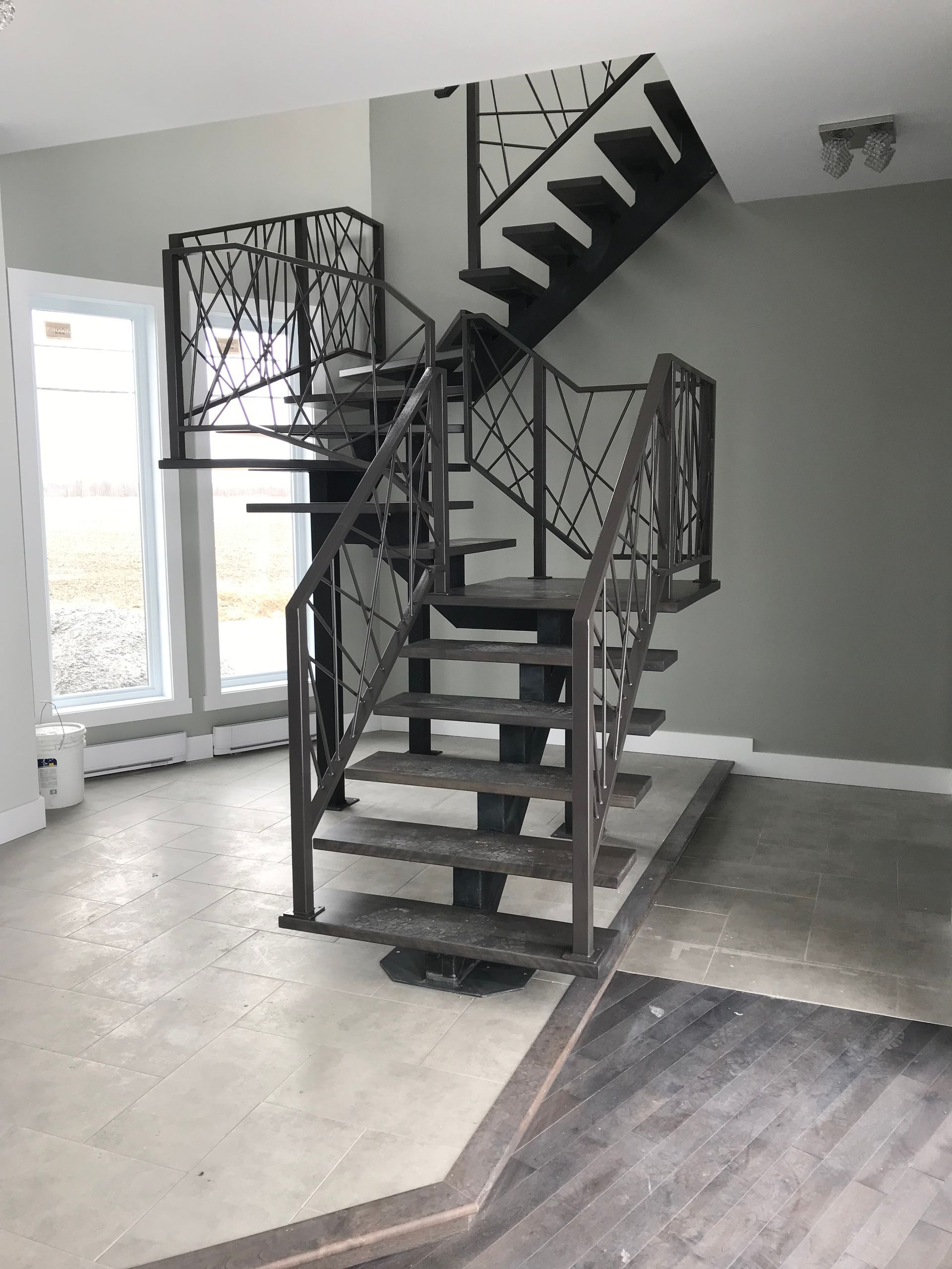Escalier métallique avec rampes ouvragées, murs gris et lumière naturelle provenant d'une fenêtre.