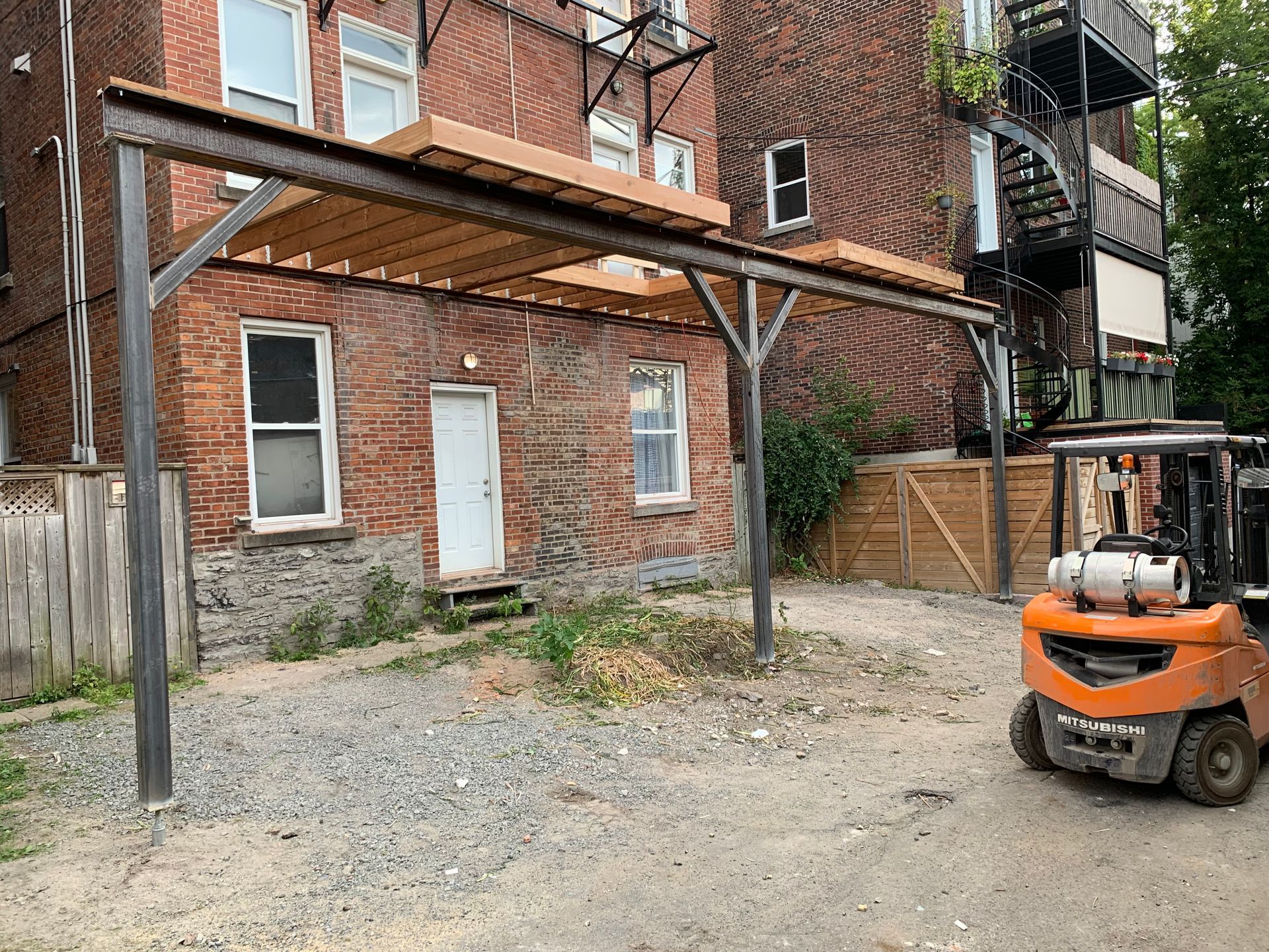 Une structure métallique abrite un auvent au-dessus d'une petite cour. Un chariot élévateur est stationné à droite, près d'un bâtiment en briques.