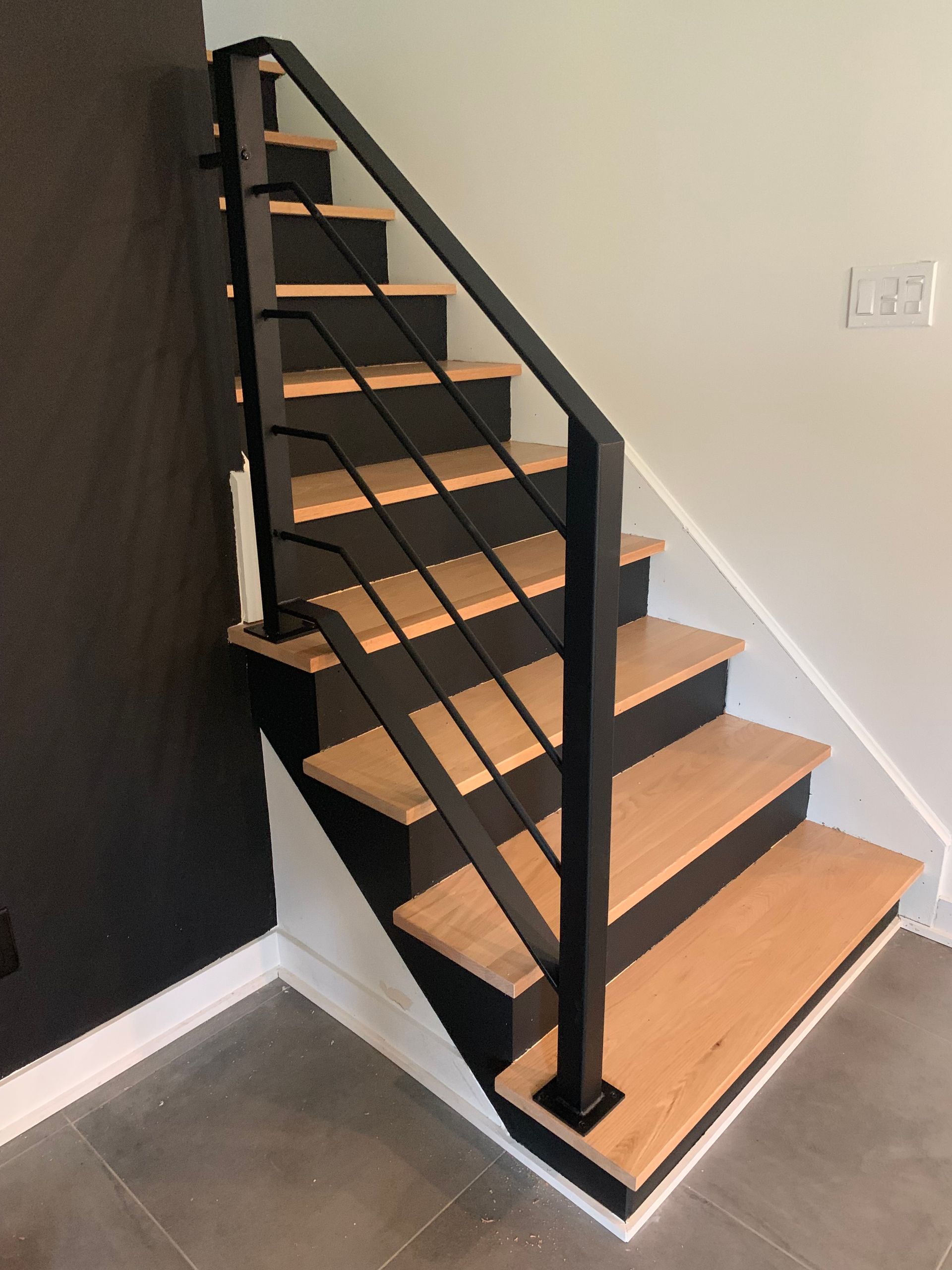 Escalier avec marches en bois et rampe en métal noir ; mur foncé à gauche et mur clair à droite.