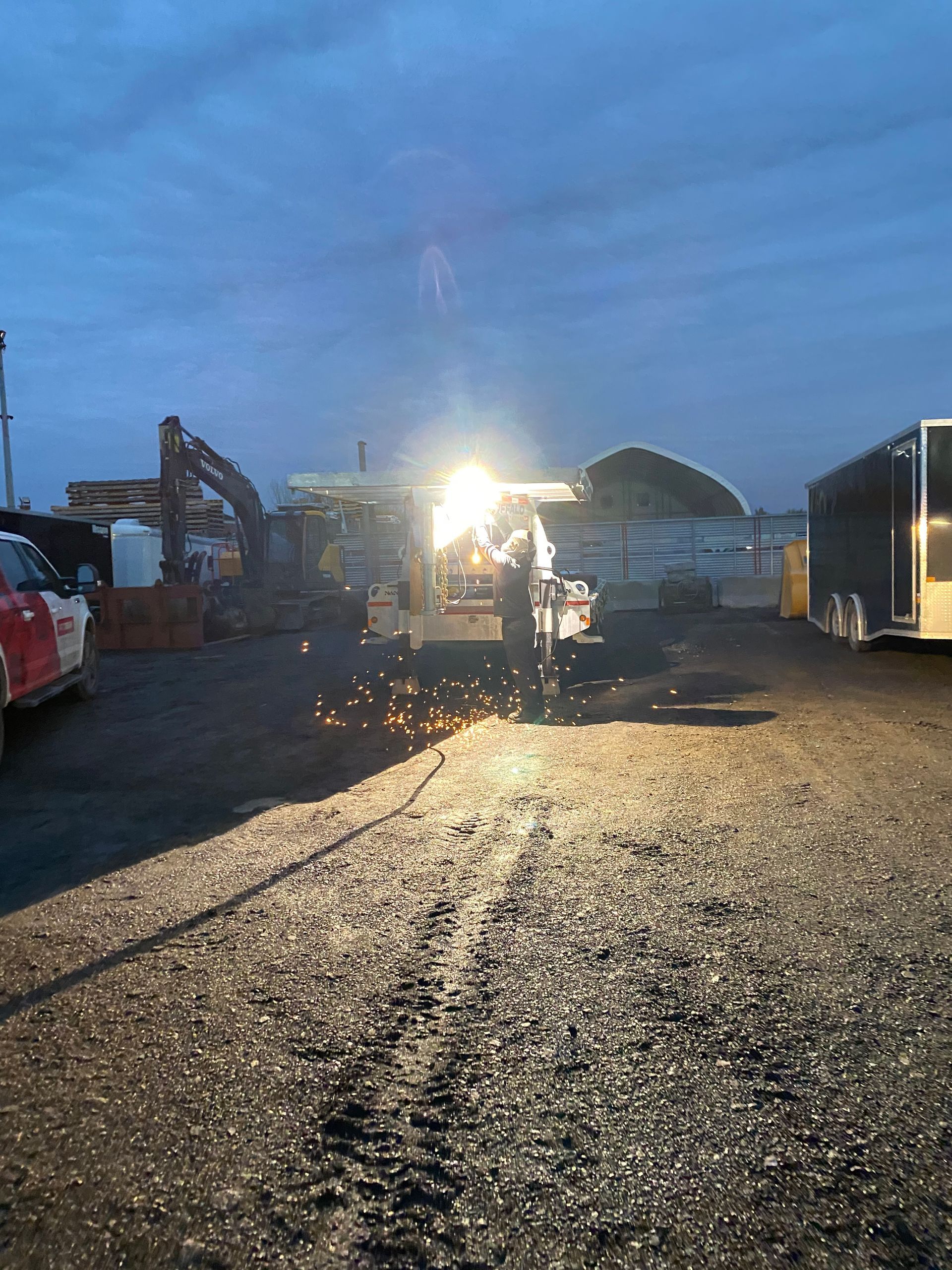 Un soudeur travaille sur du métal, des étincelles jaillissent, près d'une excavatrice, d'une remorque et sous un ciel crépusculaire.