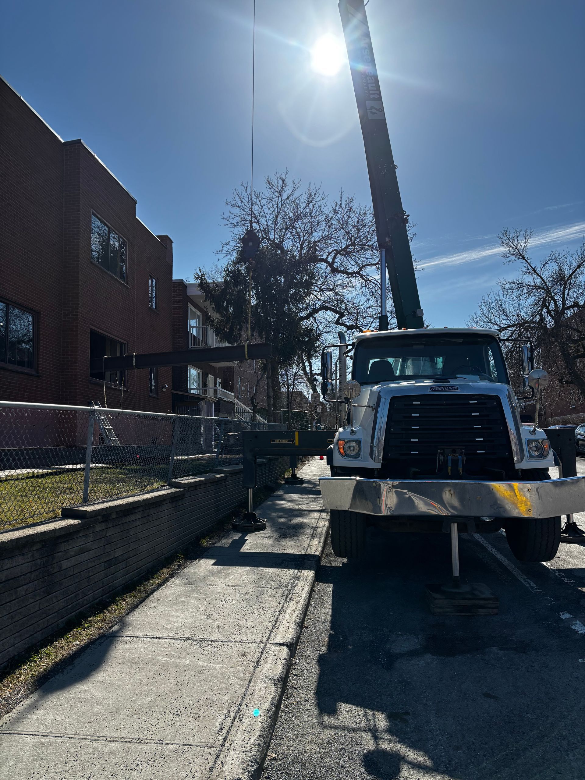 Camion-grue dans une rue de la ville ; levage d'une poutre métallique près d'un bâtiment en briques par une journée ensoleillée.