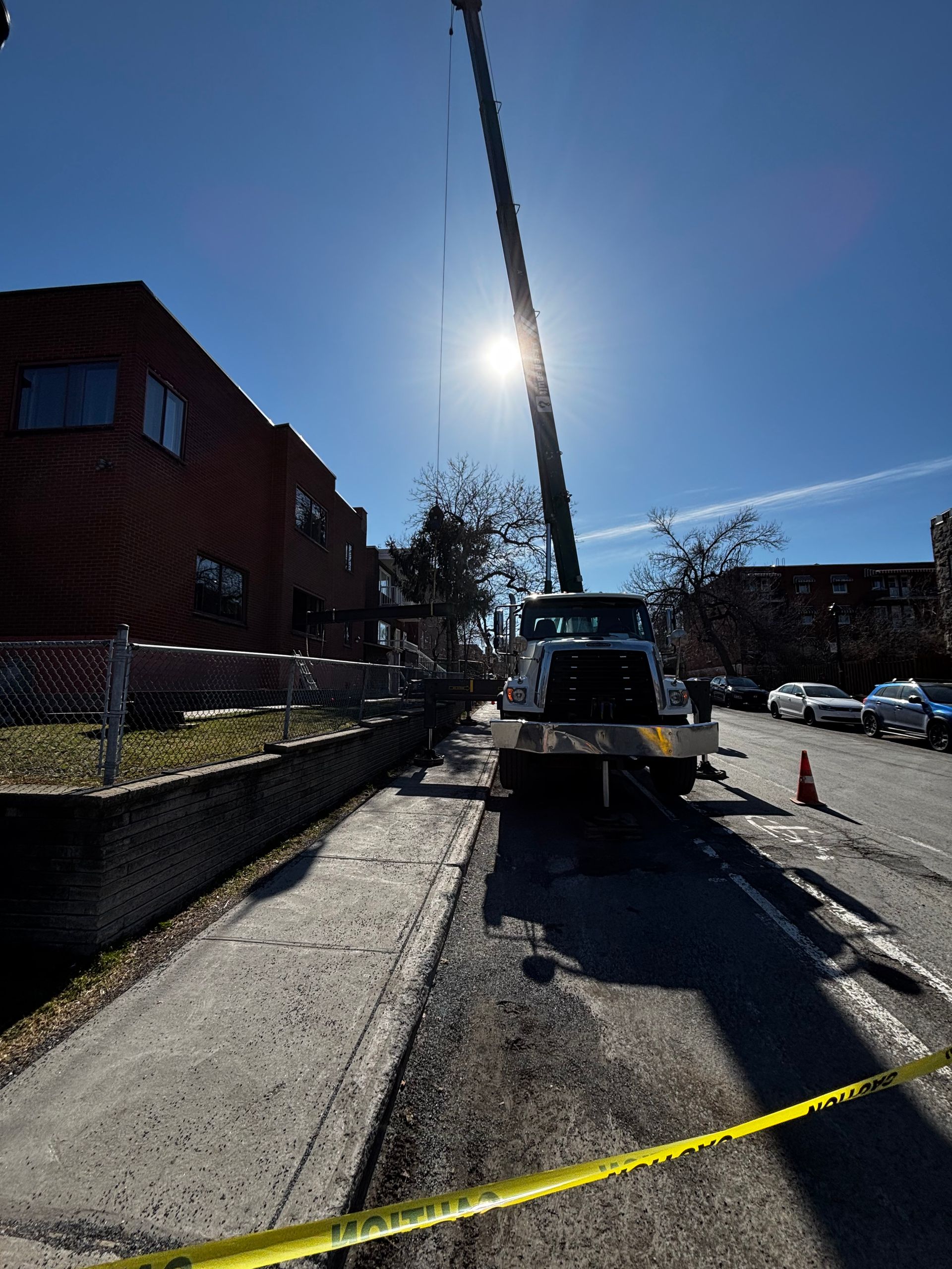 Un camion-grue est stationné dans la rue, probablement en train de démonter ou d'installer quelque chose. On aperçoit des bâtiments et un ciel ensoleillé.