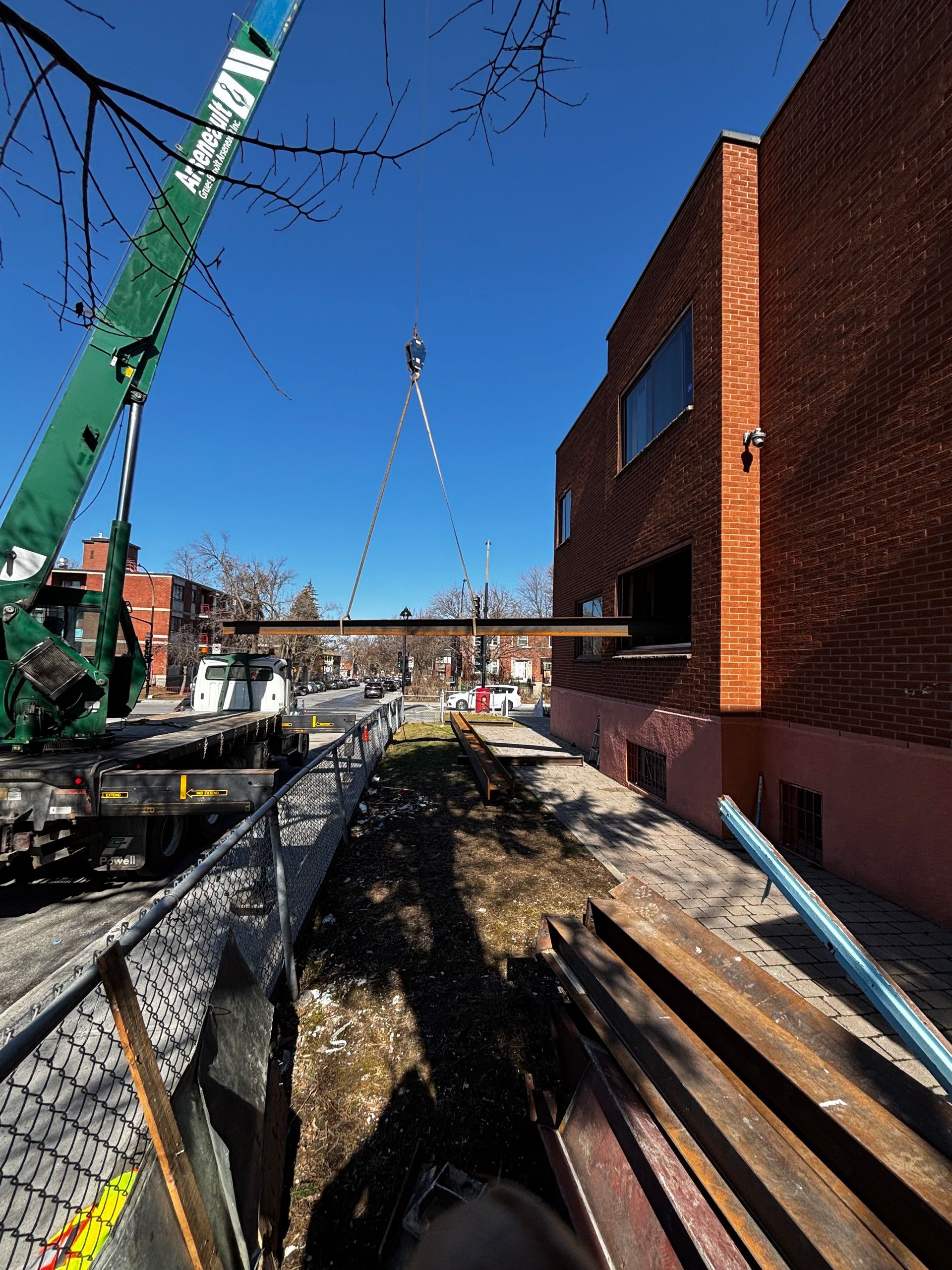 Une grue soulève une longue poutre métallique vers un bâtiment dont une fenêtre nouvellement créée s'ouvre par une journée ensoleillée.