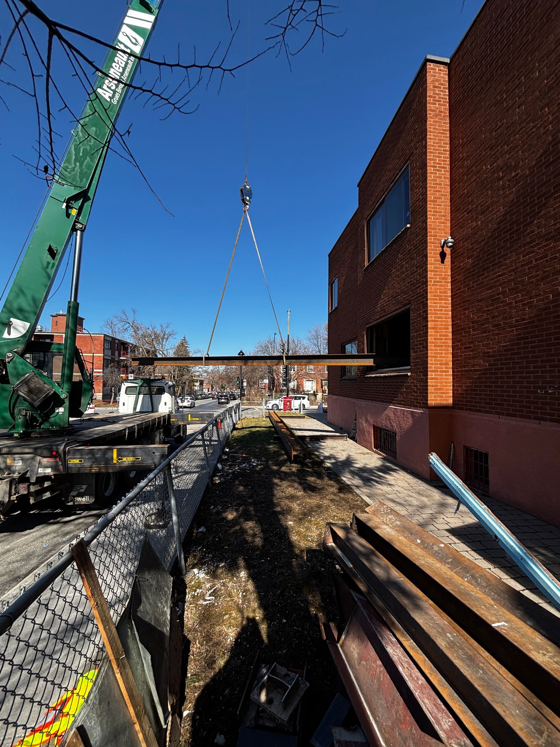 Une grue soulève une longue poutre d'acier près d'un bâtiment en briques par une journée ensoleillée.