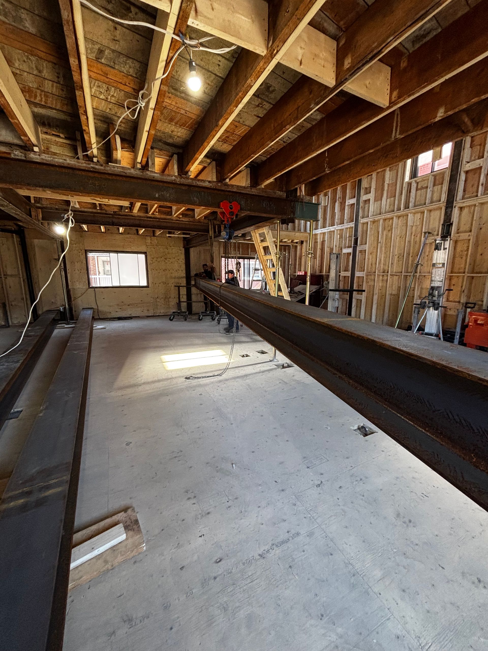 Intérieur d'un chantier de construction : sol en béton, poutres en bois apparentes, poutres en acier et ossature murale partielle.