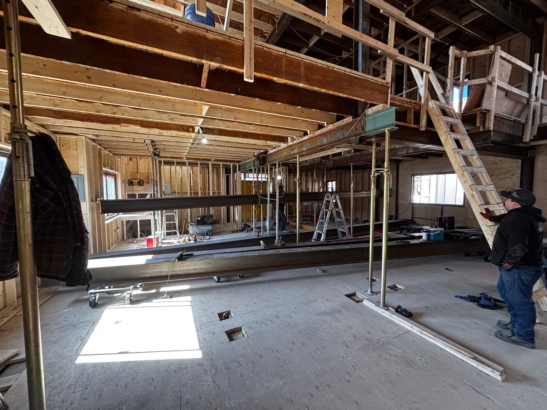 Intérieur d'un chantier de construction avec poutres apparentes, échafaudages et une personne coiffée d'un chapeau inspectant l'avancement des travaux.