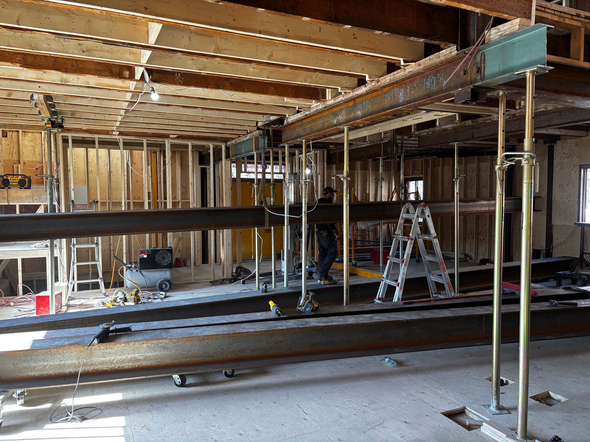 Vue intérieure d'un chantier de construction montrant des poutres d'acier, une charpente en bois, des échafaudages et un ouvrier.