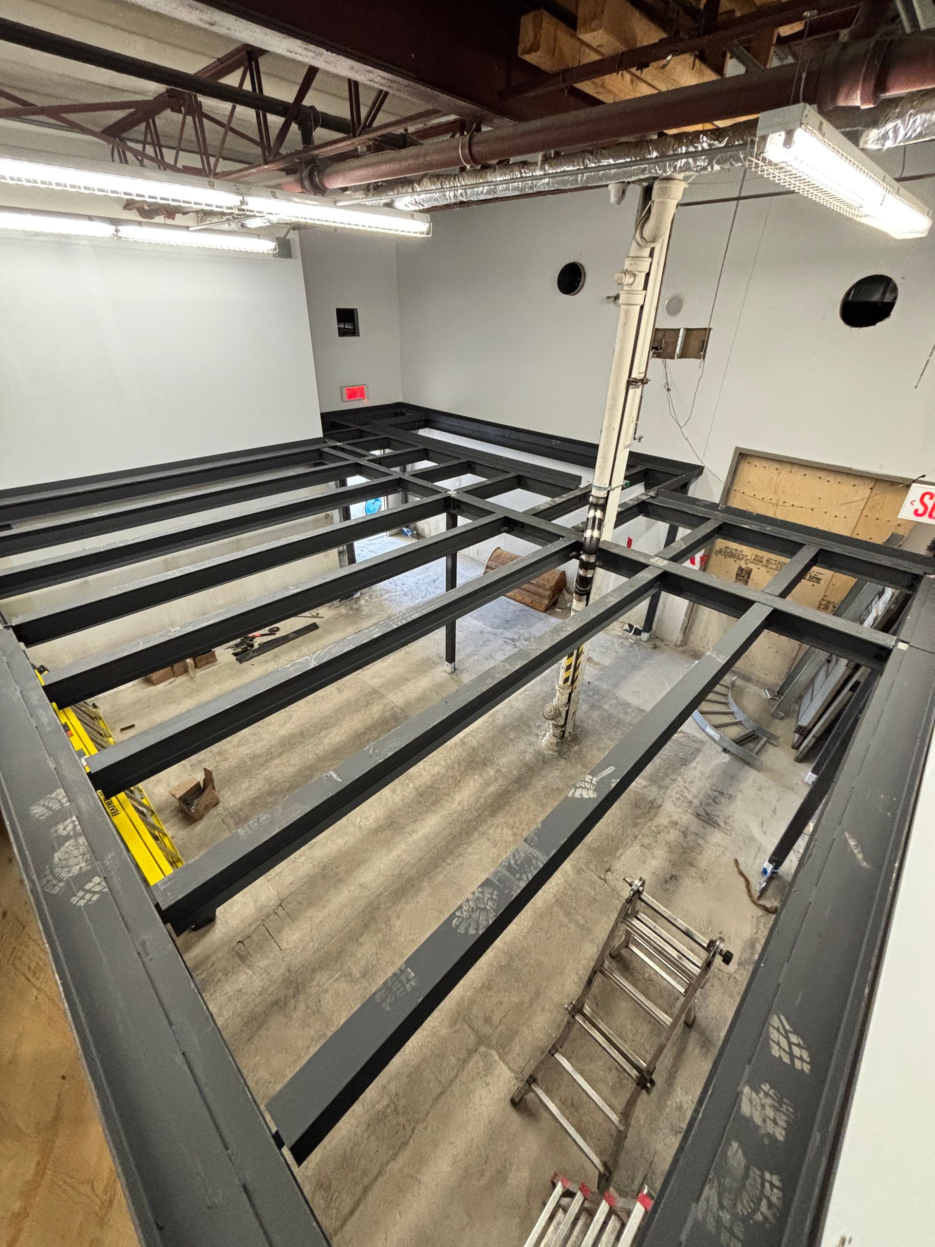 Vue aérienne d'une pièce à la structure en acier noir, probablement un deuxième étage ou une plateforme. Murs blancs, éclairage fluorescent.