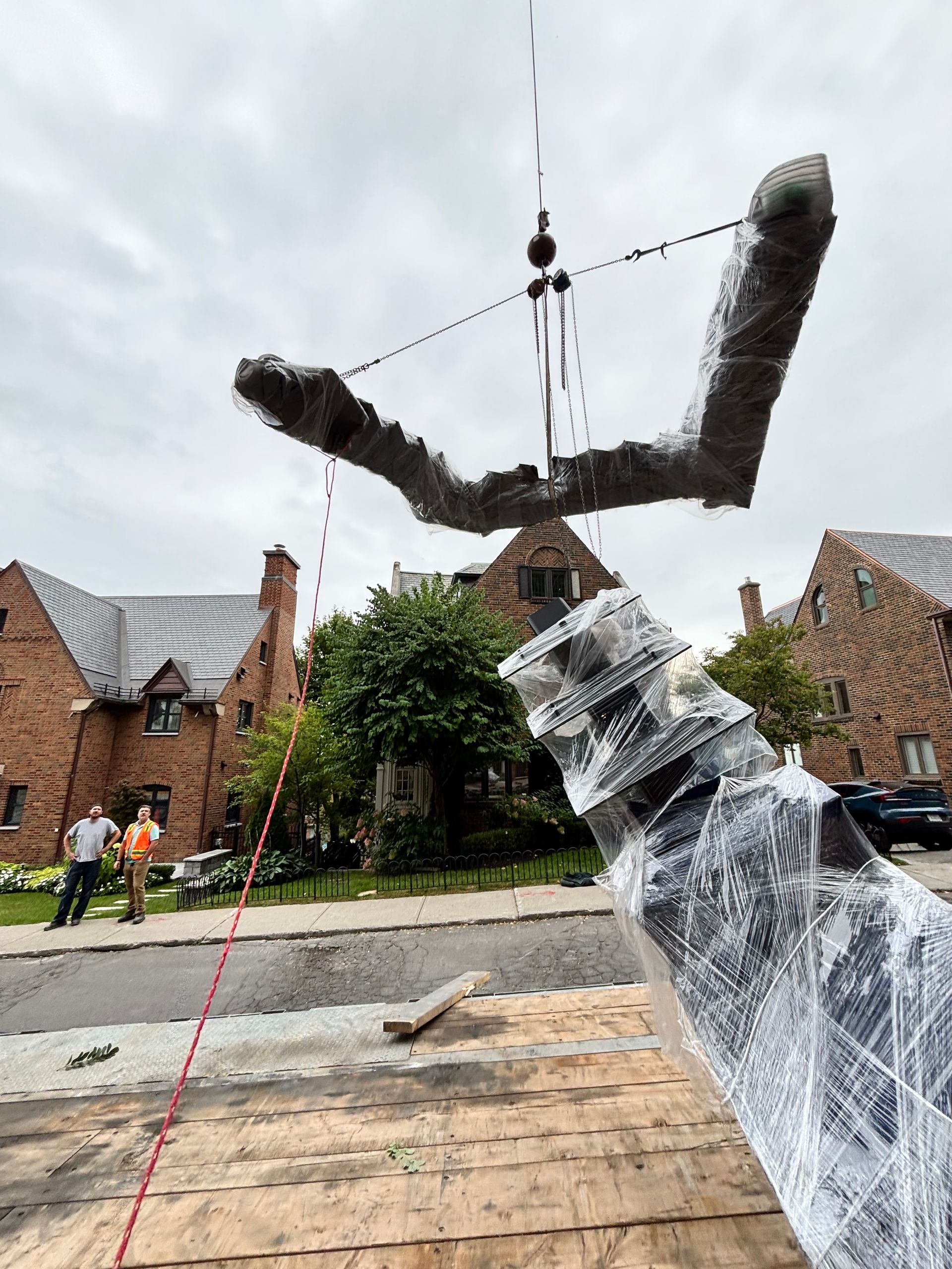 Une grue soulève un grand élément sculptural emballé au-dessus d'une rue bordée de maisons en briques.