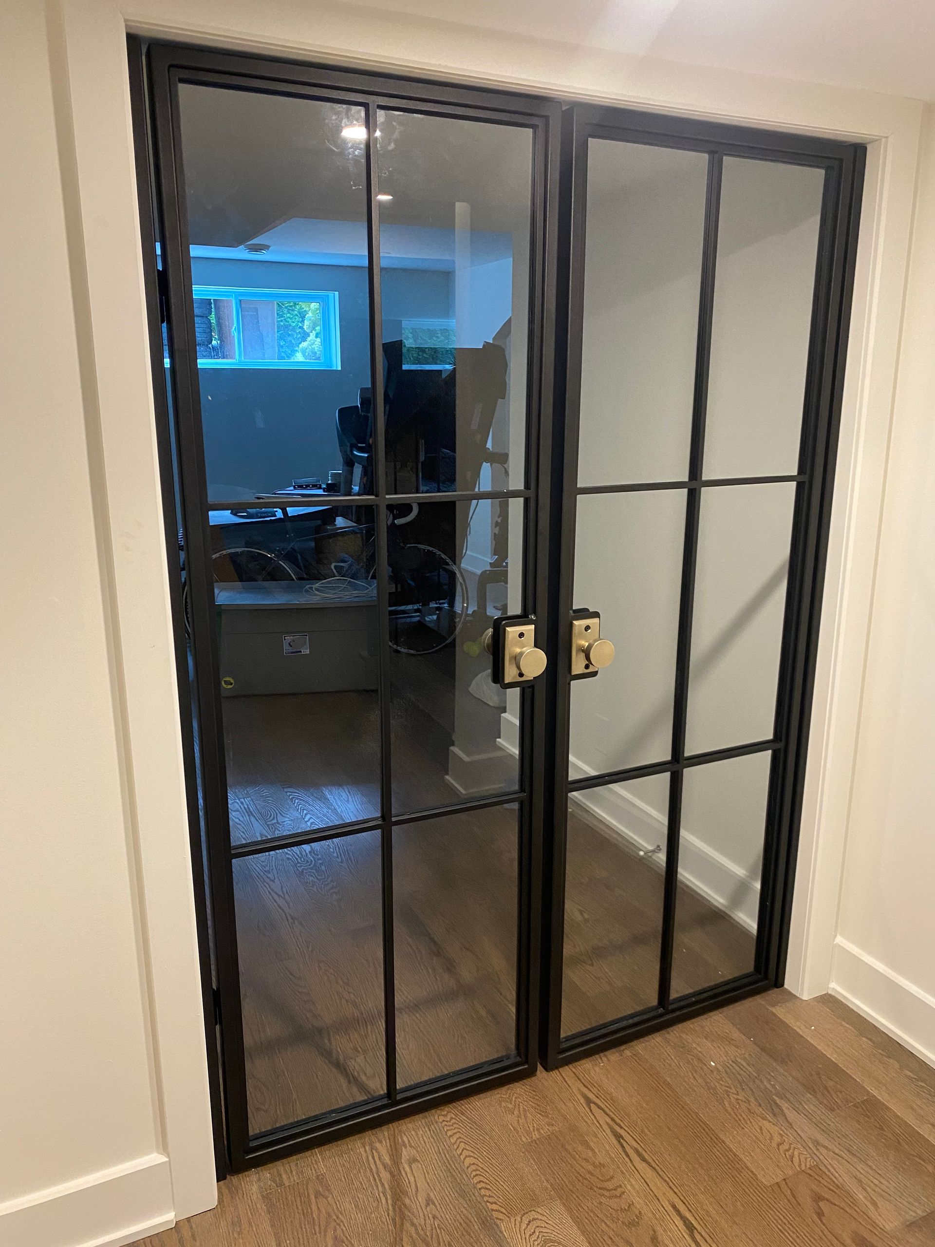 Portes vitrées à cadre noir et motif quadrillé, quincaillerie en laiton, installées dans une entrée aux murs blancs ; parquet visible.