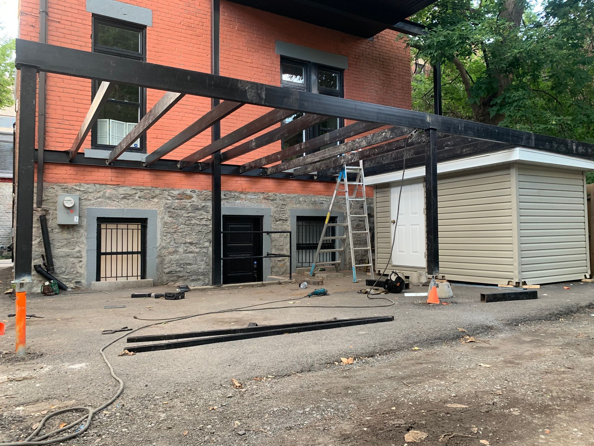 Construction d'une pergola noire adossée à un bâtiment en briques. Une échelle se trouve à proximité.