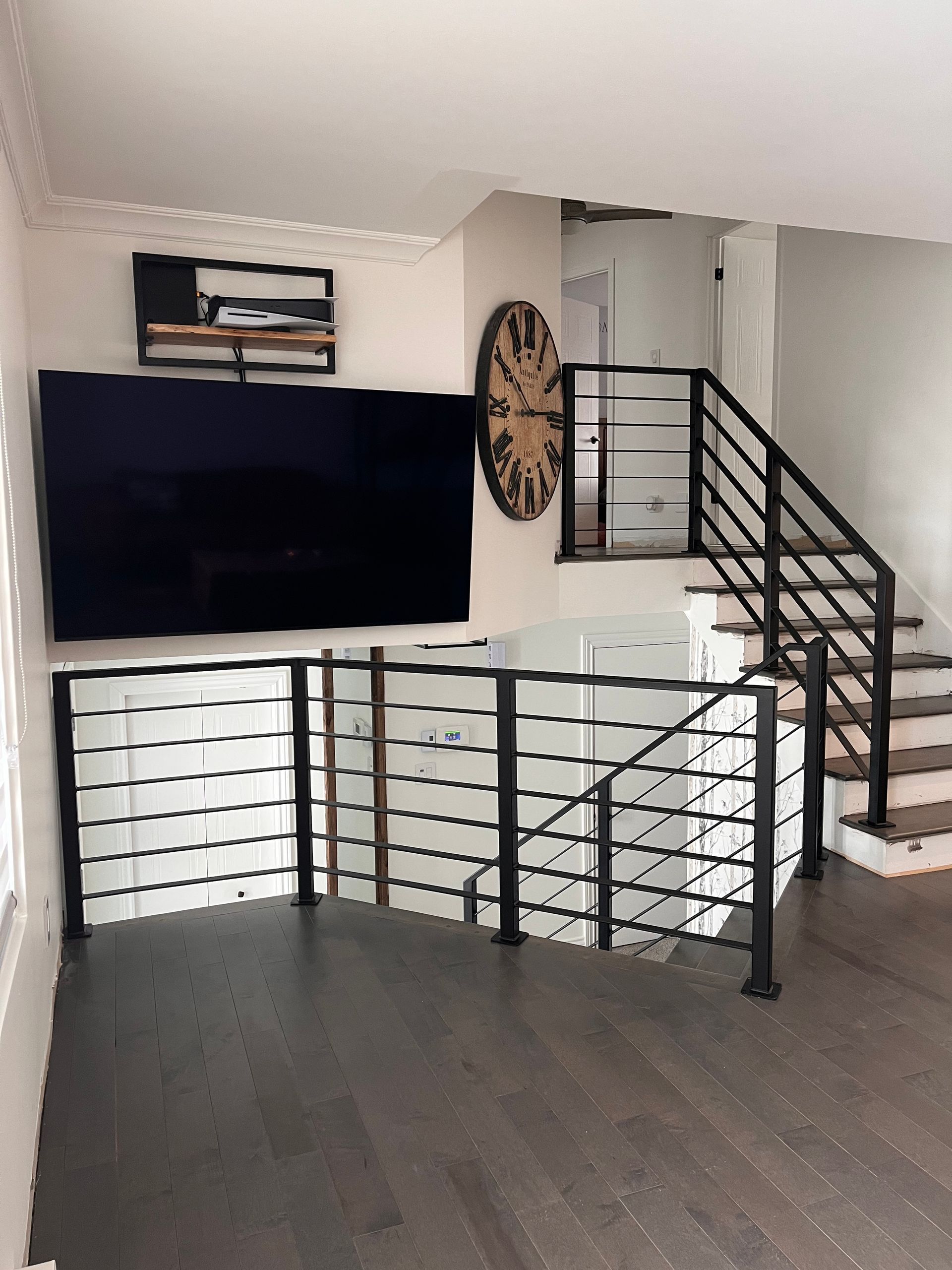 Espace de vie avec parquet foncé, escalier, rampe noire, télévision fixée au mur et horloge murale décorative.