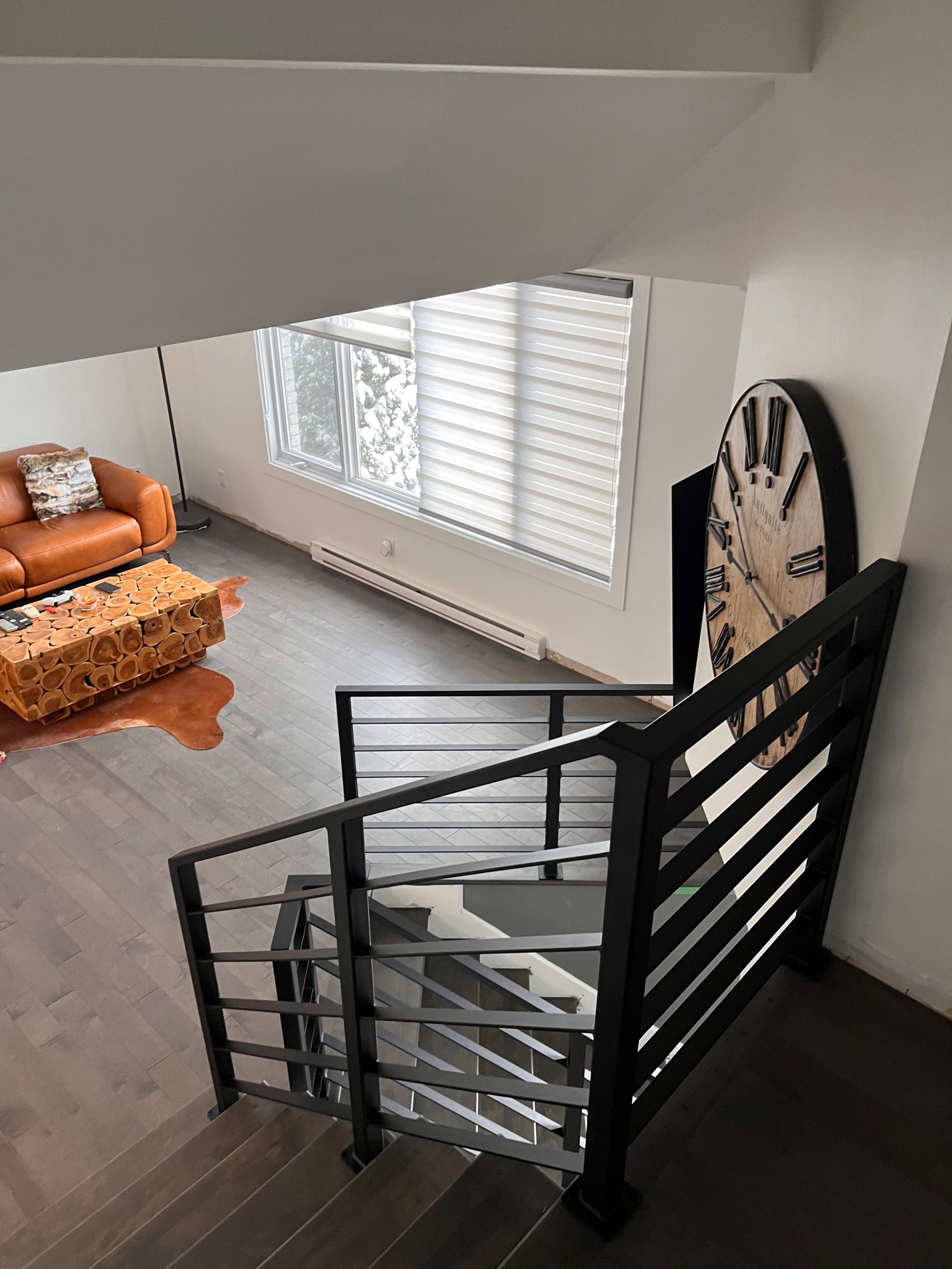 Vue depuis l'escalier : Intérieur moderne avec parquet, rampe noire, horloge surdimensionnée, canapé en cuir et stores.