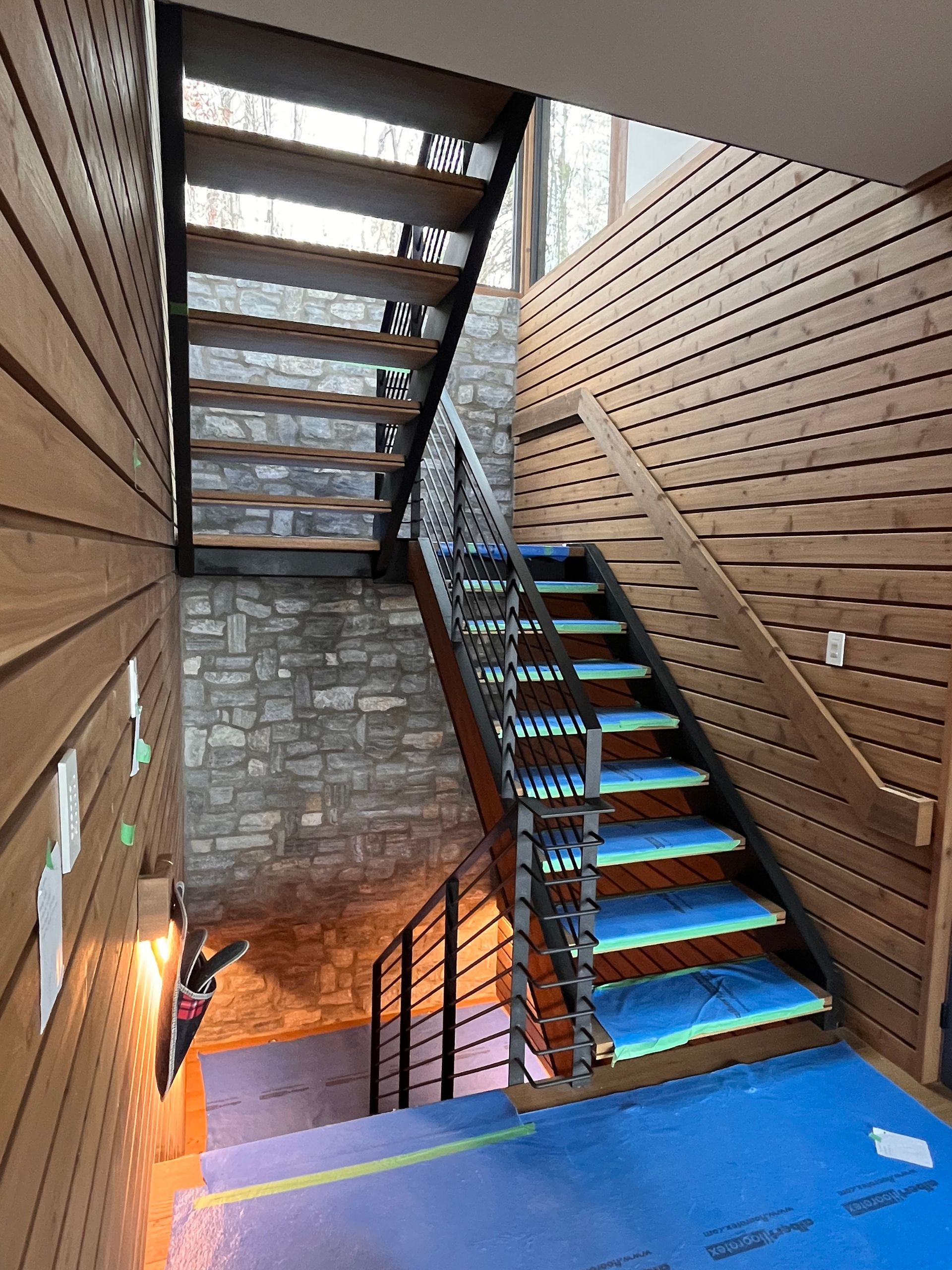 Escalier avec marches en bois et rampes en métal, situé entre des murs en bois et en pierre ; revêtement protecteur bleu sur les marches inférieures.
