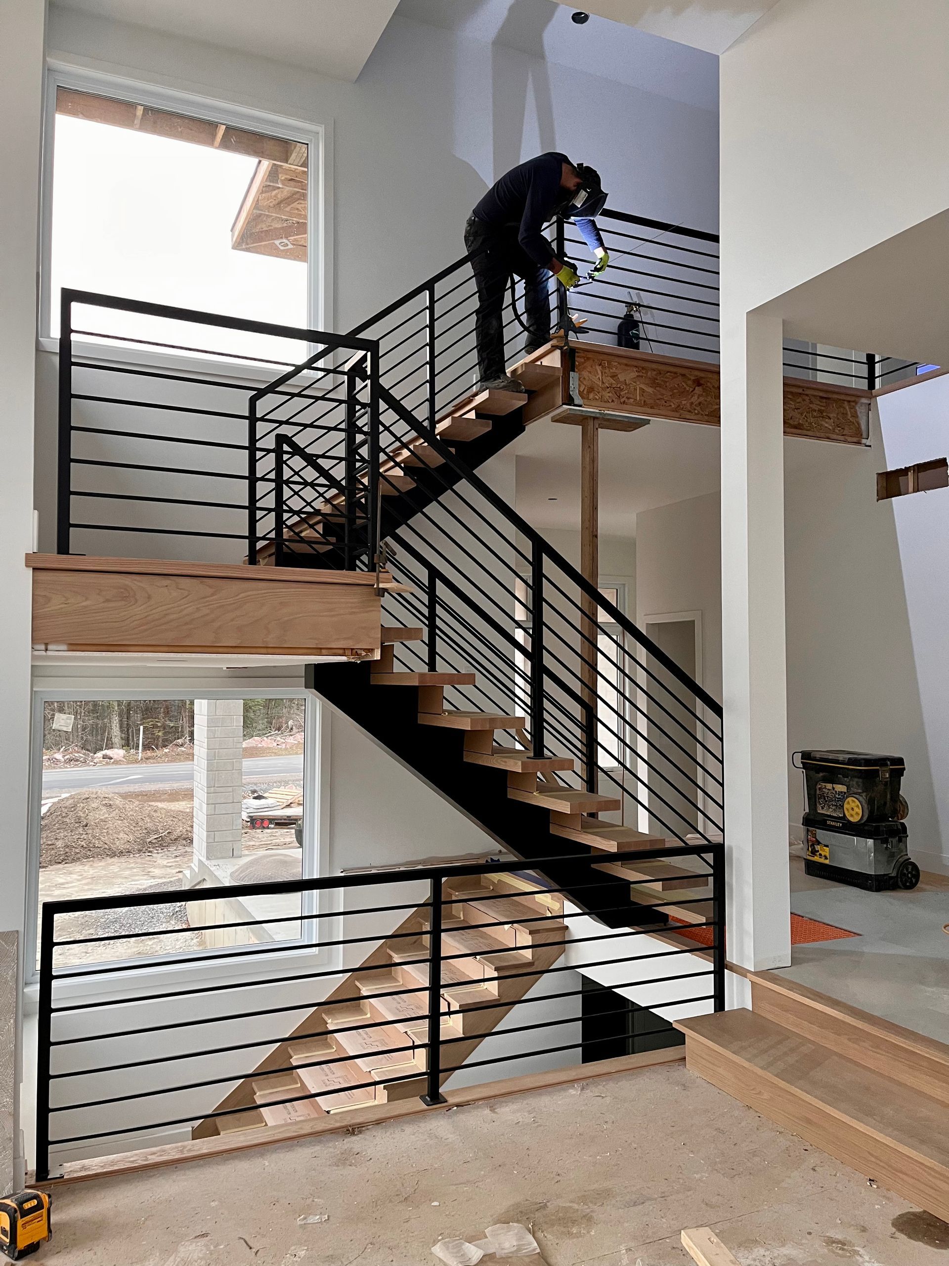 Une personne travaille sur un escalier moderne aux rampes en métal noir. Marches en bois clair, murs blancs et grande fenêtre.