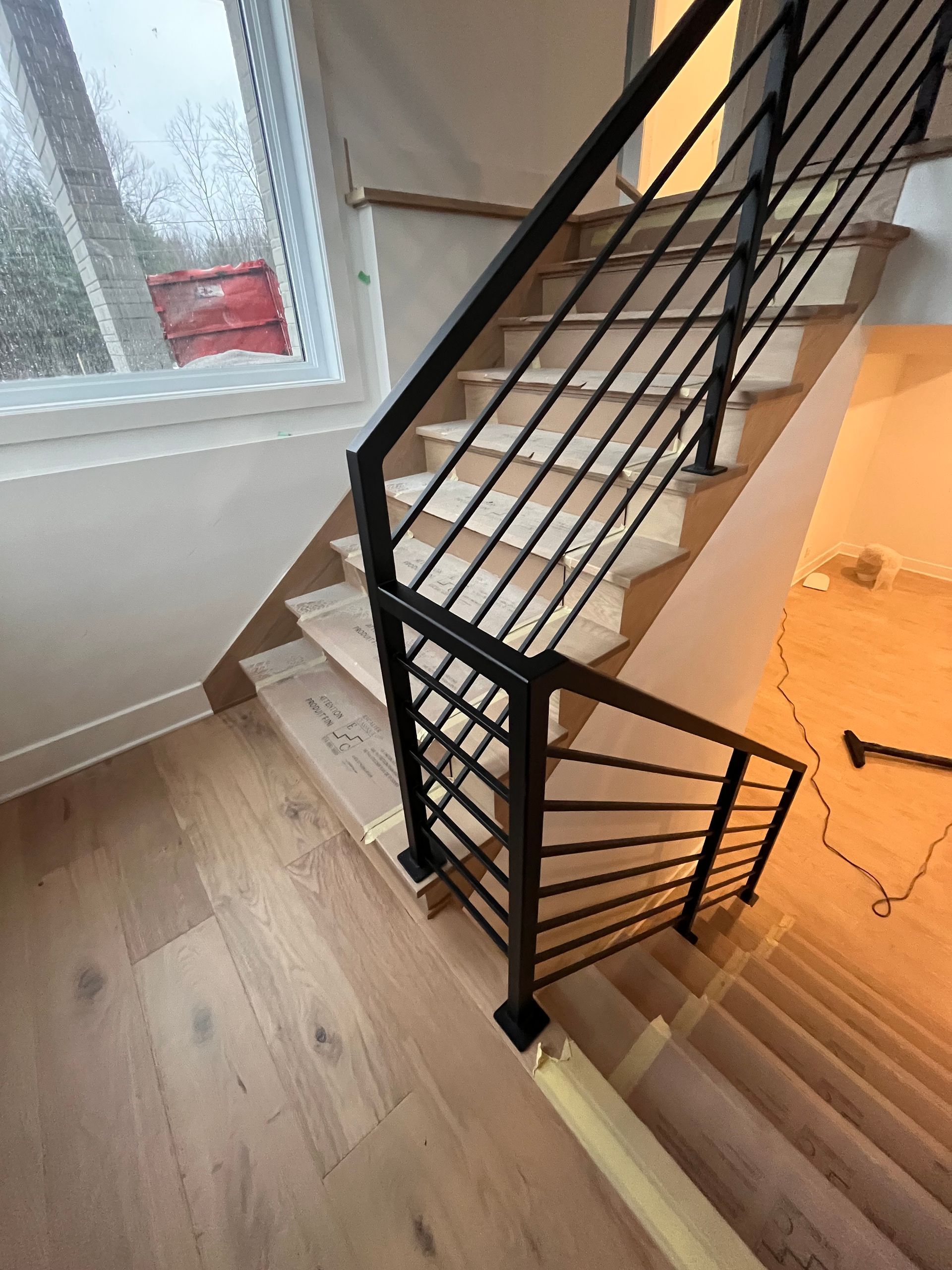 Escalier avec marches en bois et rampes en métal noir, menant à l'étage. Fenêtre en arrière-plan.