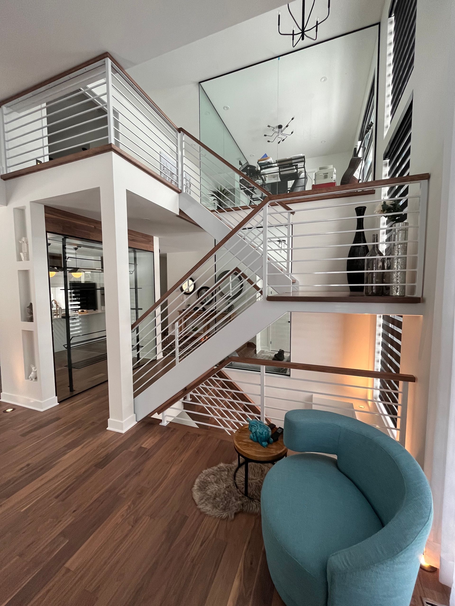 Intérieur sur deux étages avec parquet, escalier, rampes blanches et fauteuil turquoise.
