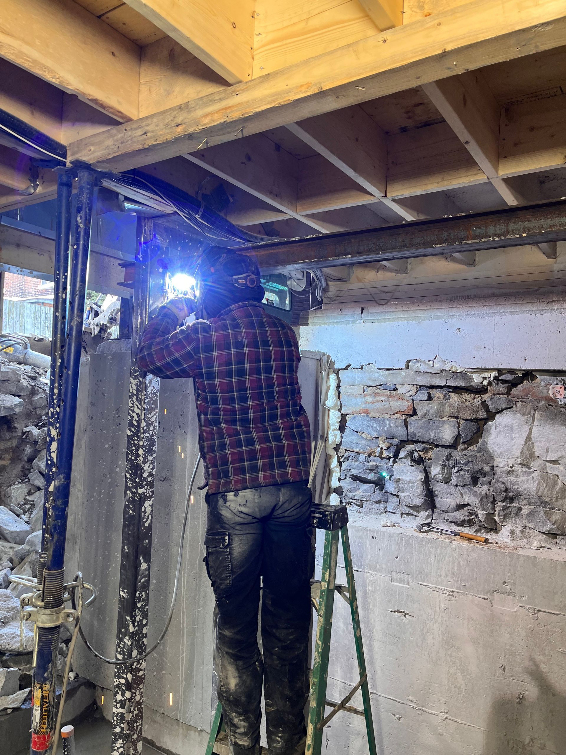 Une personne soude des poutres métalliques sous une structure en bois, debout sur un escabeau.