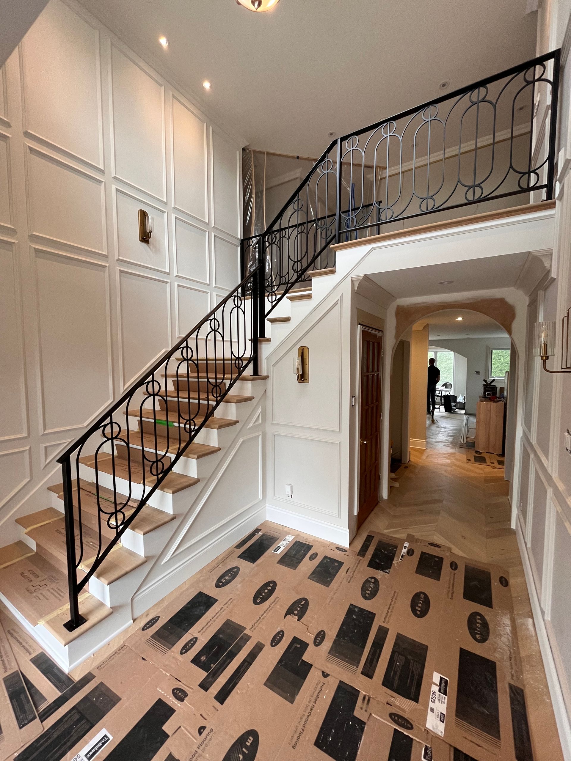 Entrée élégante avec escalier, boiseries blanches et rampe en fer forgé. Le sol est recouvert d'un revêtement protecteur.