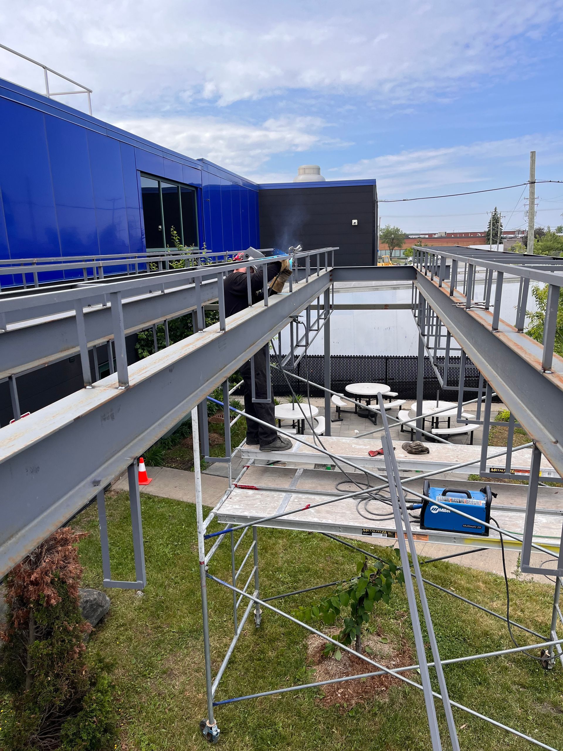 Construction d'une passerelle métallique à l'extérieur d'un bâtiment bleu, avec des échafaudages et des ouvriers visibles.