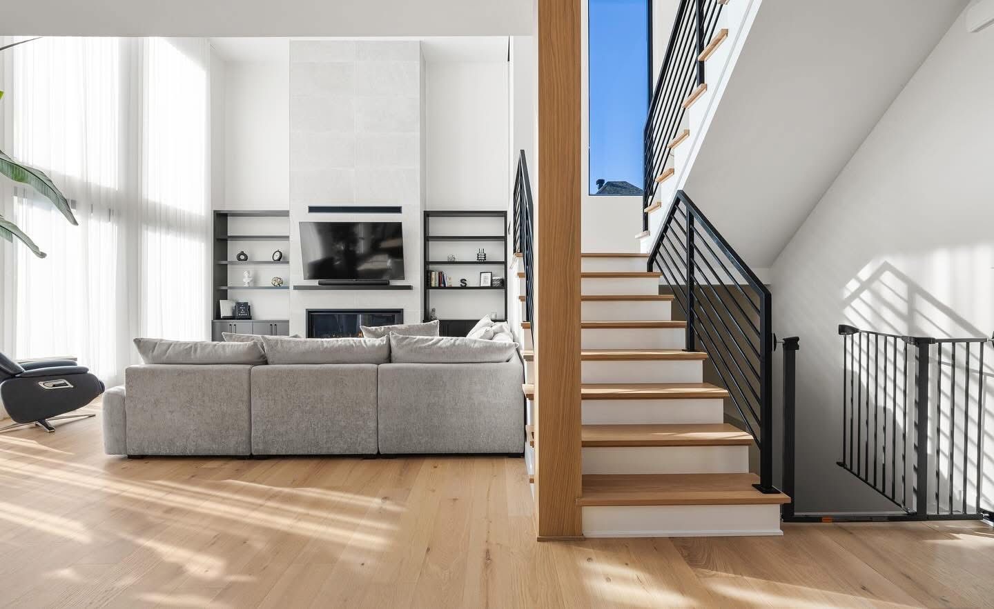 Salon avec un canapé gris clair, parquet et un escalier avec une rampe noire.