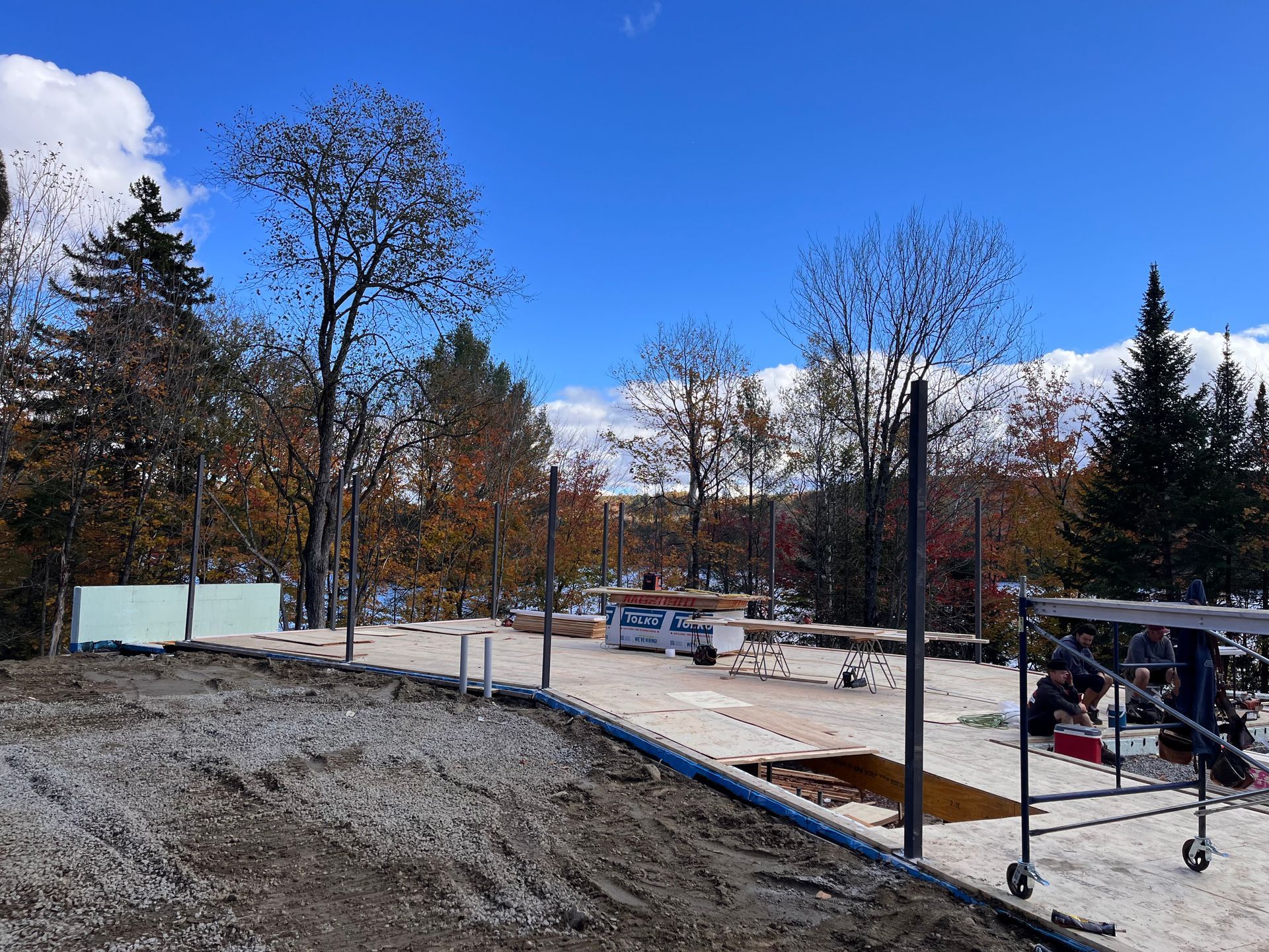 Chantier de construction avec une remorque, des poteaux et des arbres sous un ciel bleu.